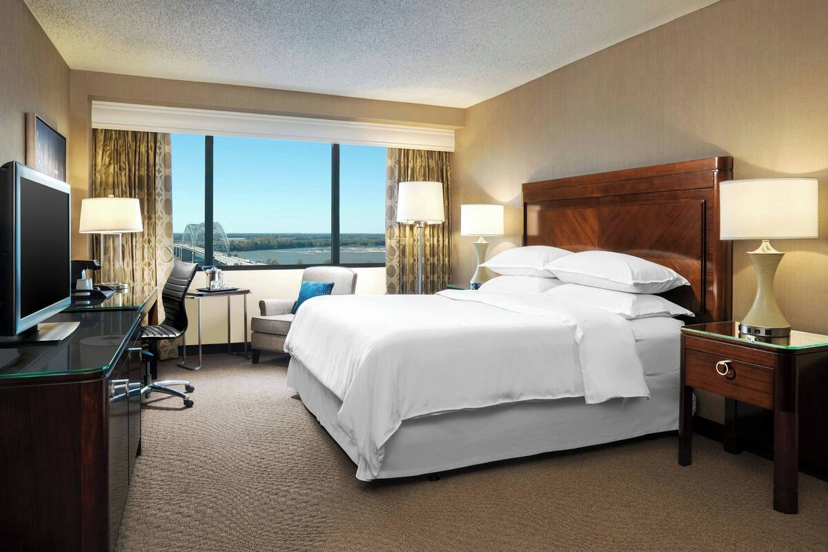 A hotel room with a large bed, a desk with a computer, a TV, and a view of a bridge and water.