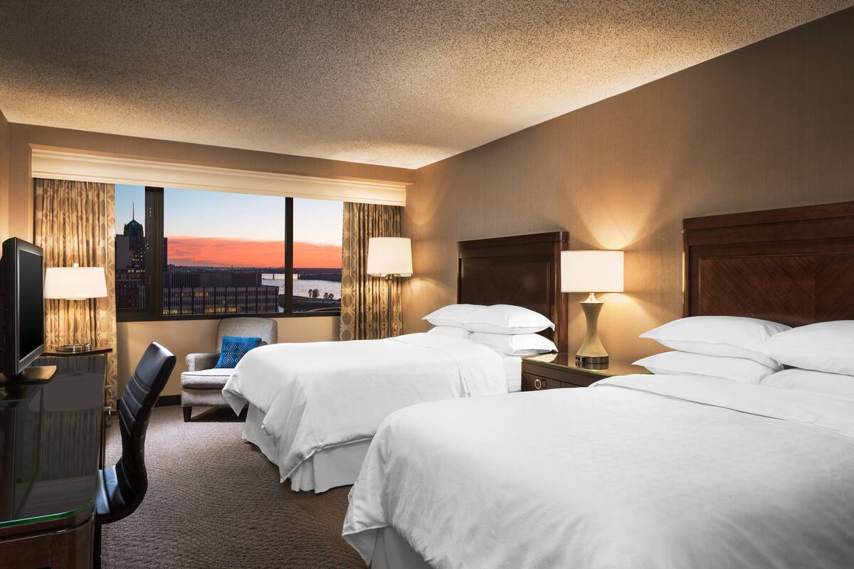 A hotel room with two beds, a desk with a chair, a TV, and a window with a cityscape view.