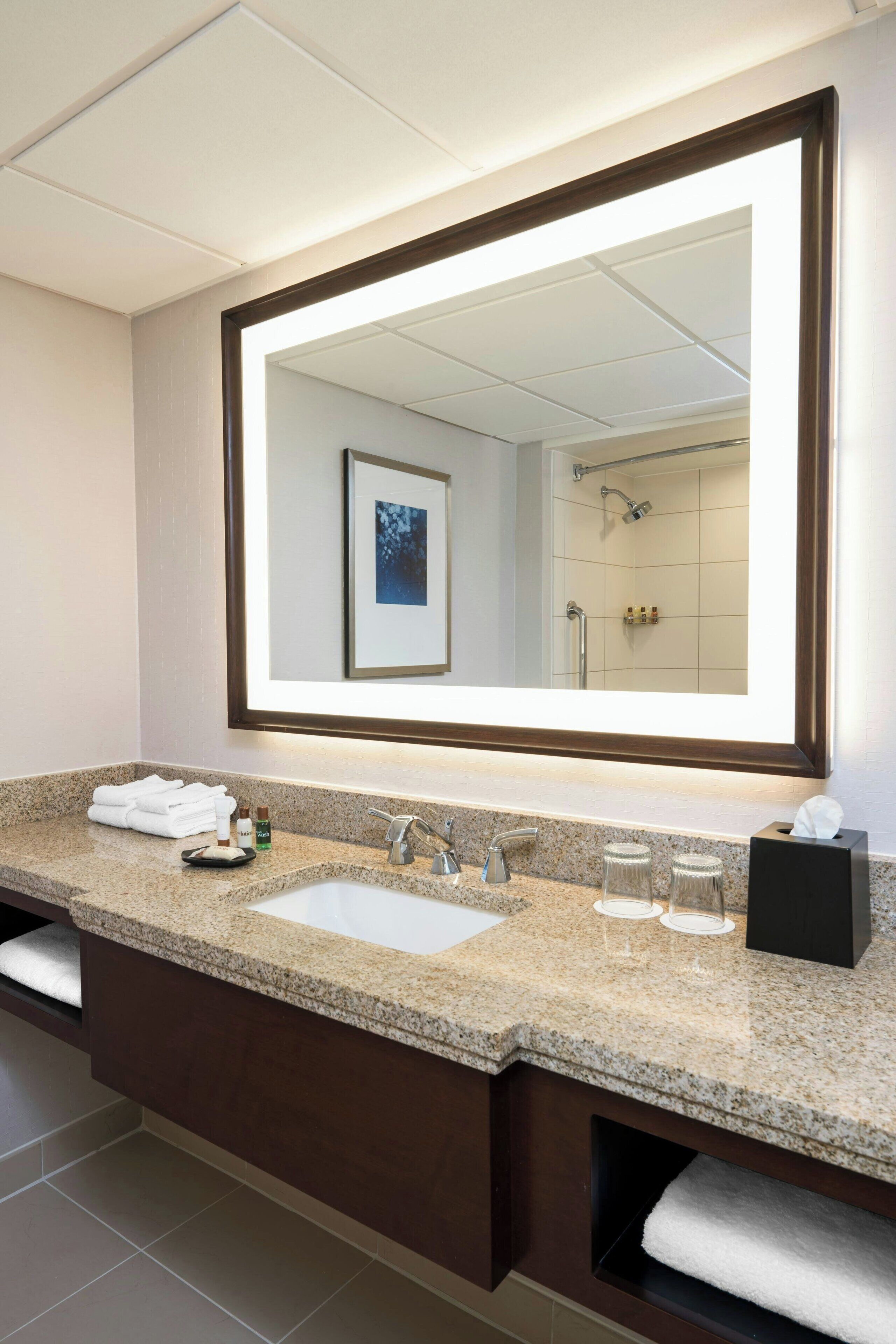 Une salle de bain moderne avec un comptoir en granit, un grand miroir, un lavabo et une zone de douche visible dans le reflet.
