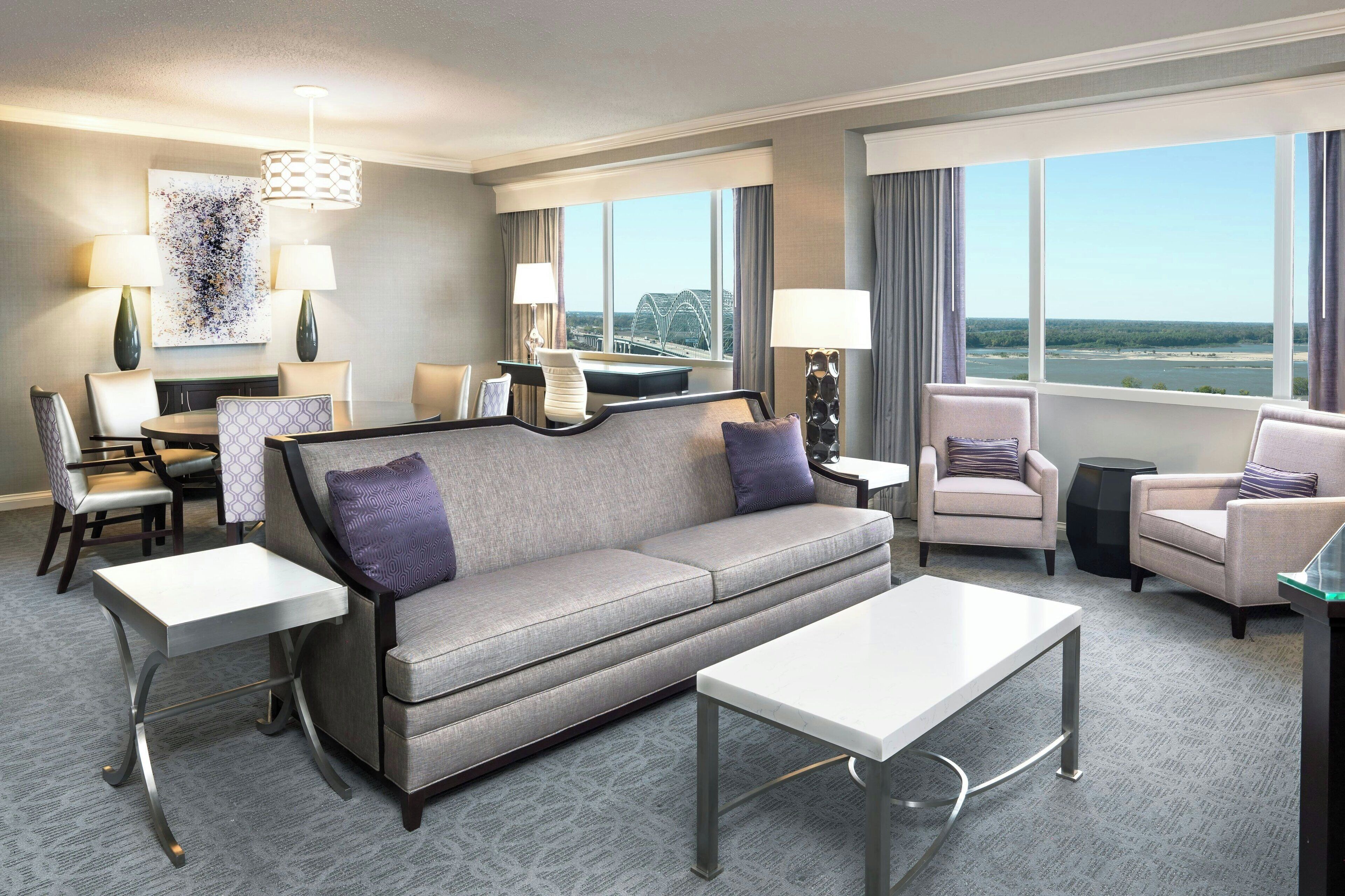 A modern hotel room with a grey sofa, two armchairs, a dining table, and a large window offering a view of a bridge and river.