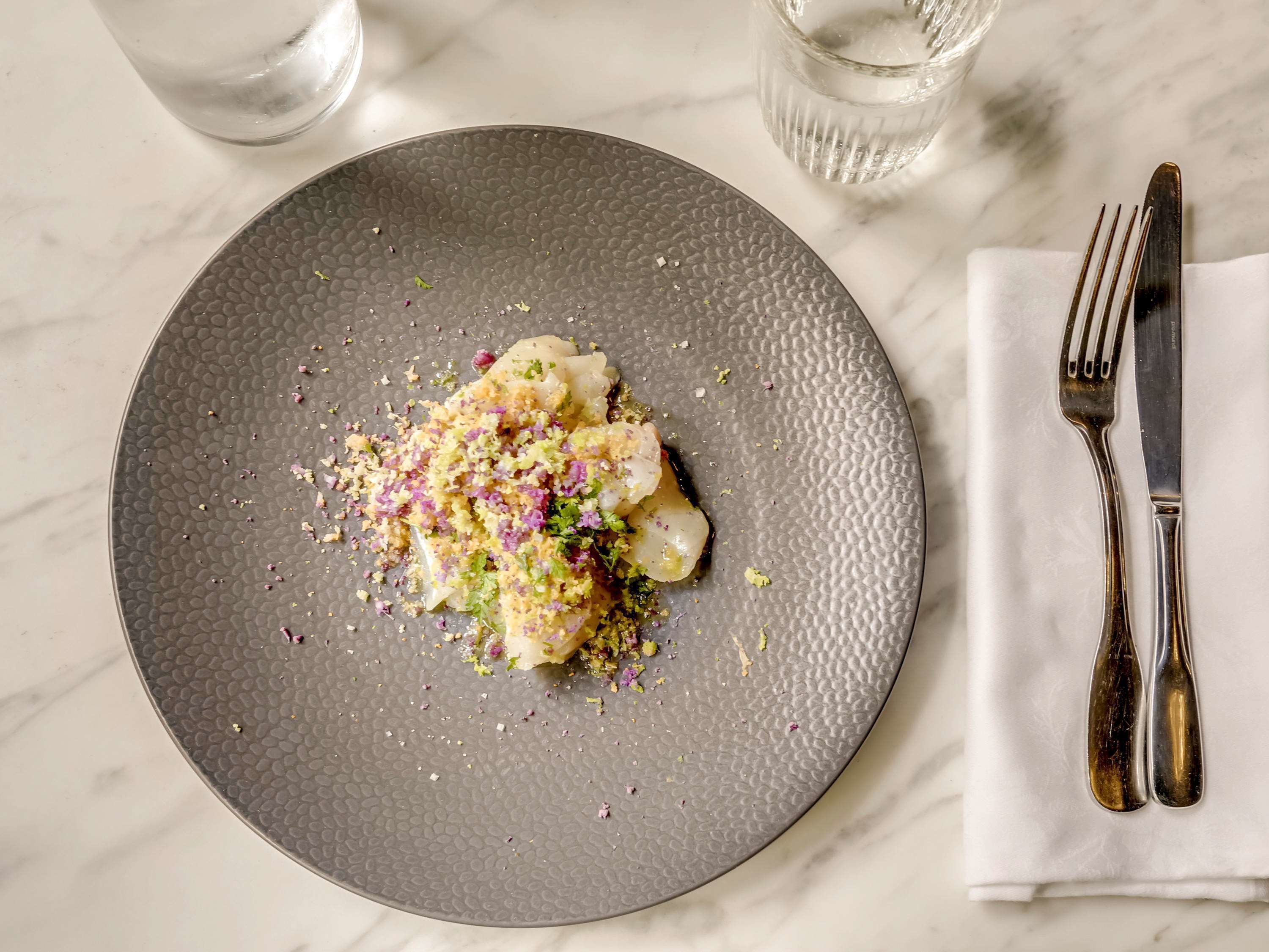 A plate of food with a textured gray plate, a fork and knife on a napkin, and two glasses of water on a marble surface.