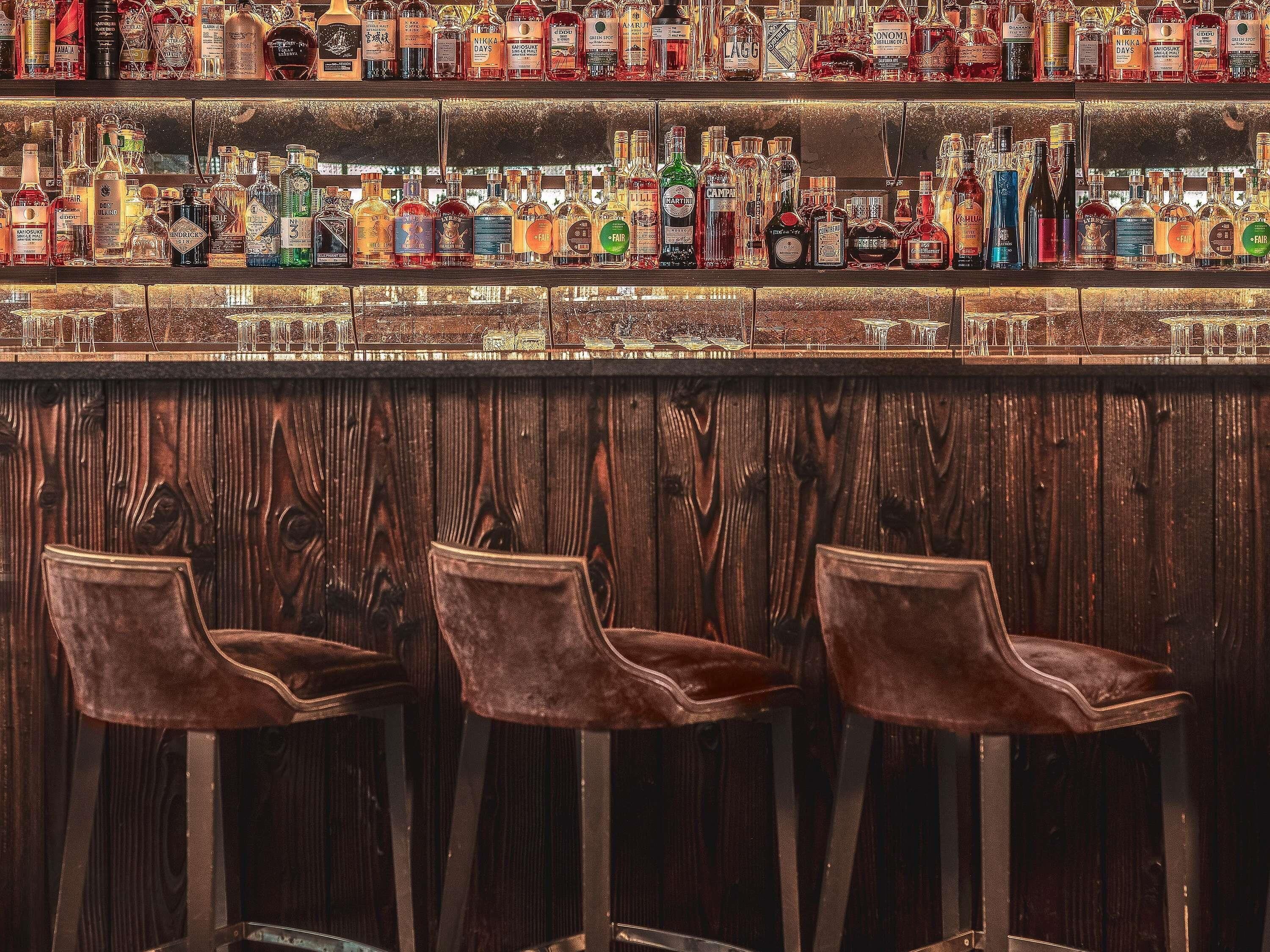 Un bar doté de plusieurs étagères remplies de bouteilles d’alcool, d’un comptoir en bois et de trois tabourets de bar marron.