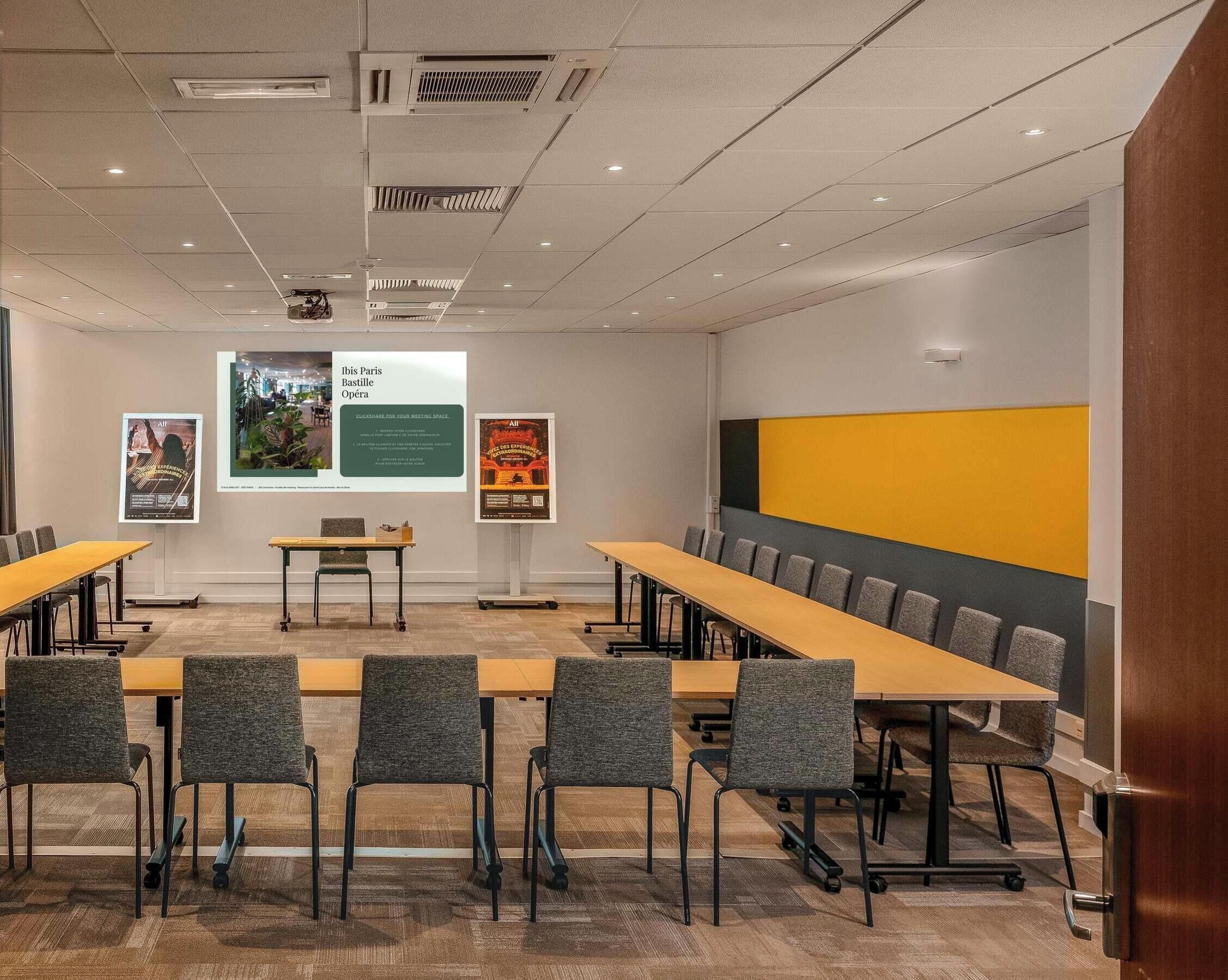 A conference room with a long table, chairs, a projector screen, and posters on the wall.