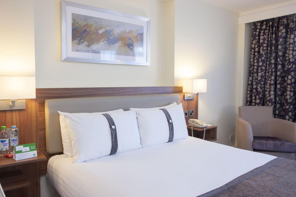 A hotel room with a large bed, two bedside lamps, a telephone, a chair, and a framed artwork on the wall.