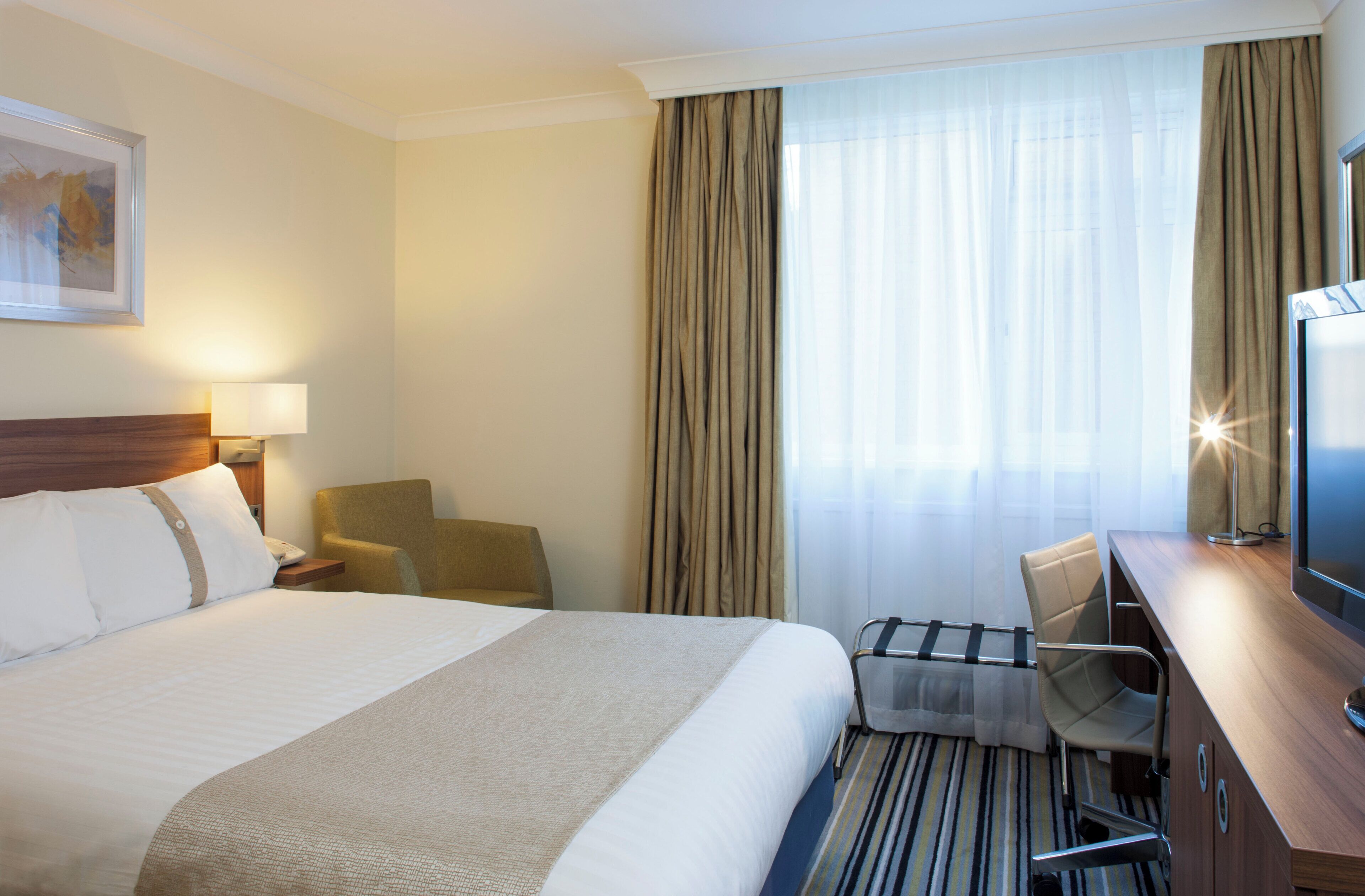Een hotelkamer met een bed, bureau, stoel, televisie en een raam met gordijnen.