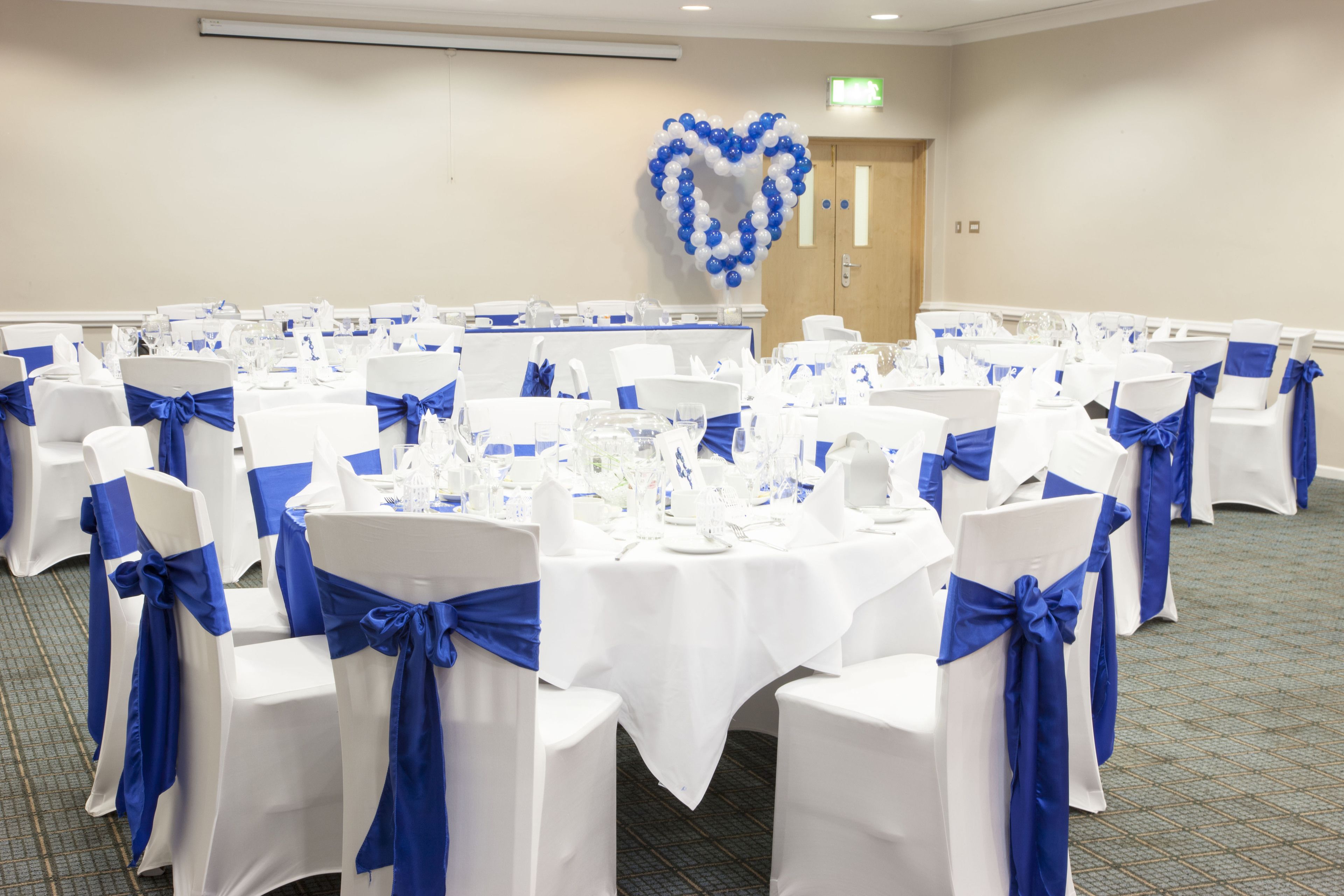 A banquet hall with round tables set for a formal event, featuring white tablecloths and blue chair sashes.