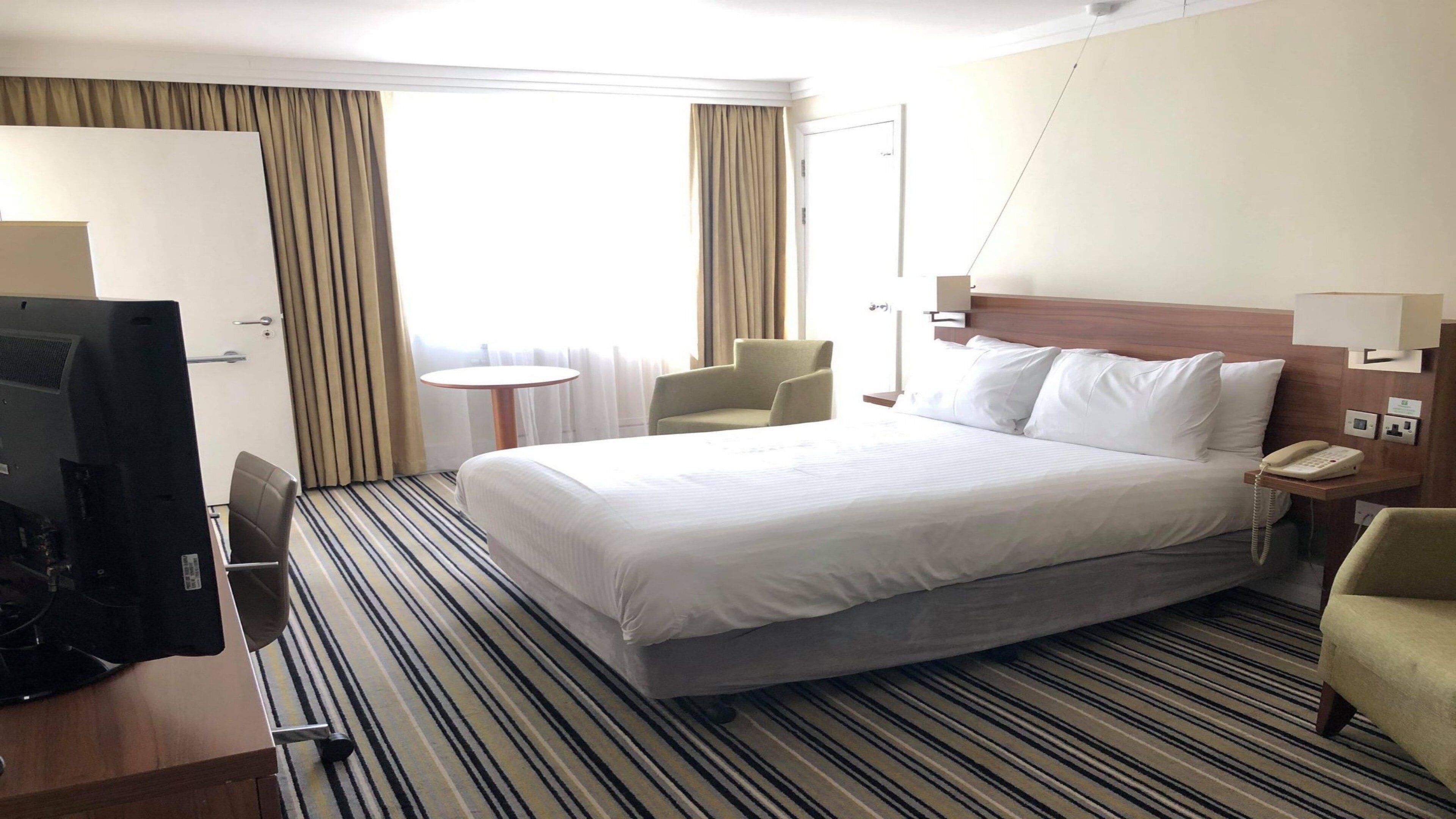 A hotel room with a large bed, a desk with a computer, a chair, a small table, and a telephone.
