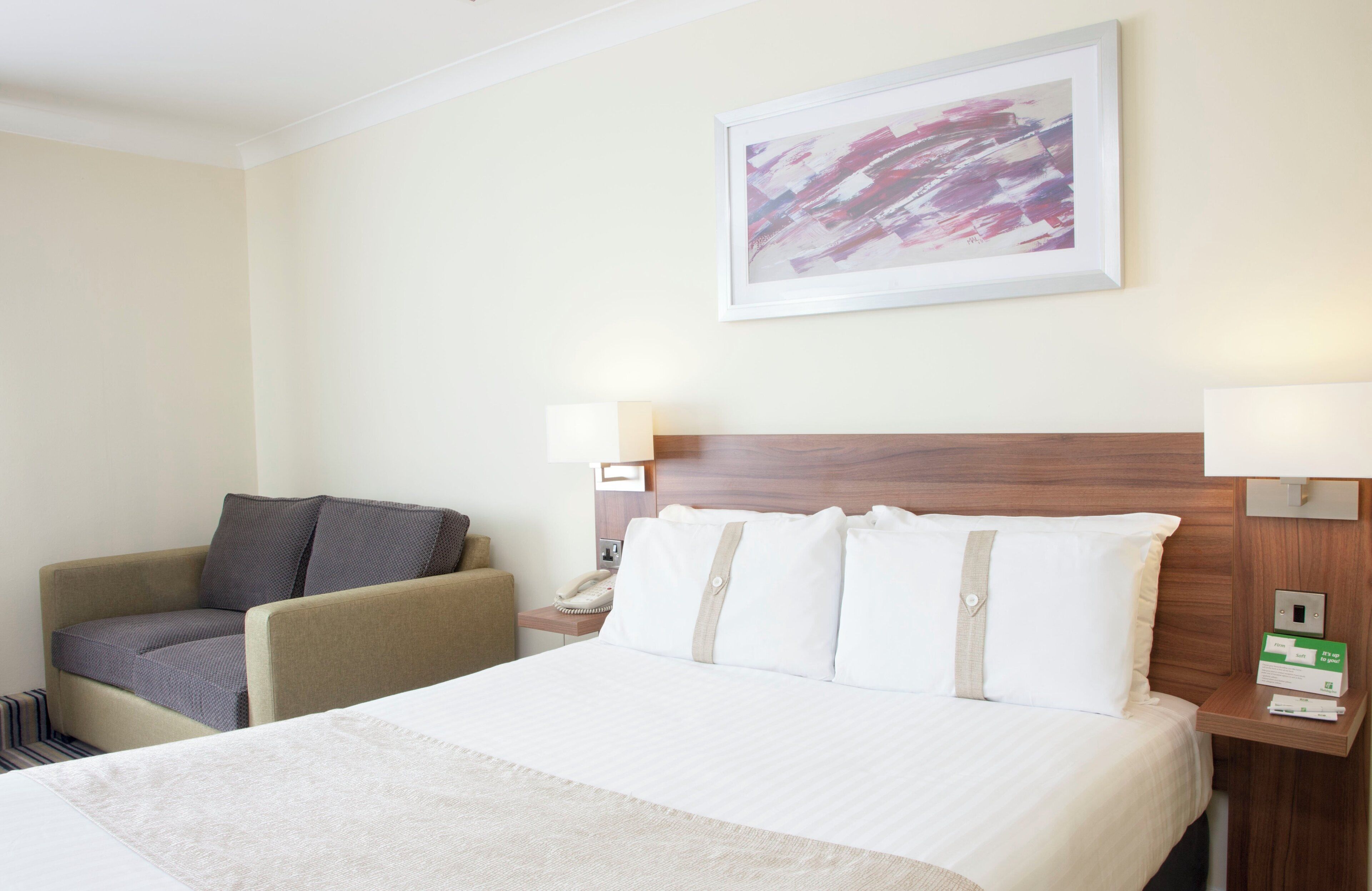 A hotel room with a bed, a chair, a nightstand with a phone, and a painting on the wall.