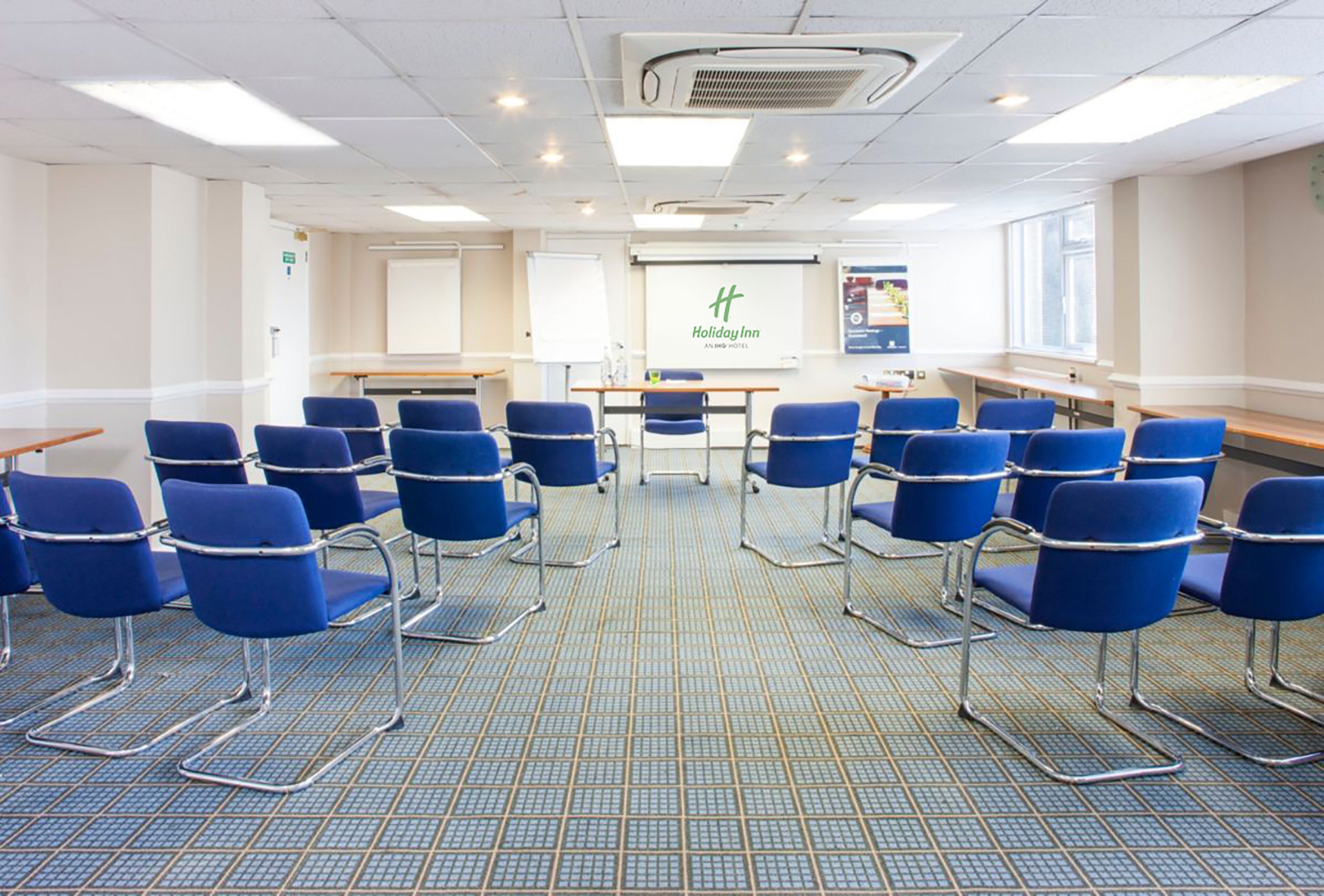 Une salle de conférence avec des chaises bleues disposées en rangées, un écran de présentation et le logo de l’hôtel Holiday Inn.