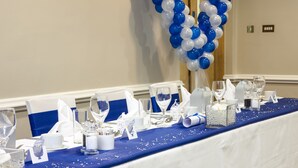 A banquet table set with white and blue decor, including a blue tablecloth, white napkins, and a blue and white balloon arch.