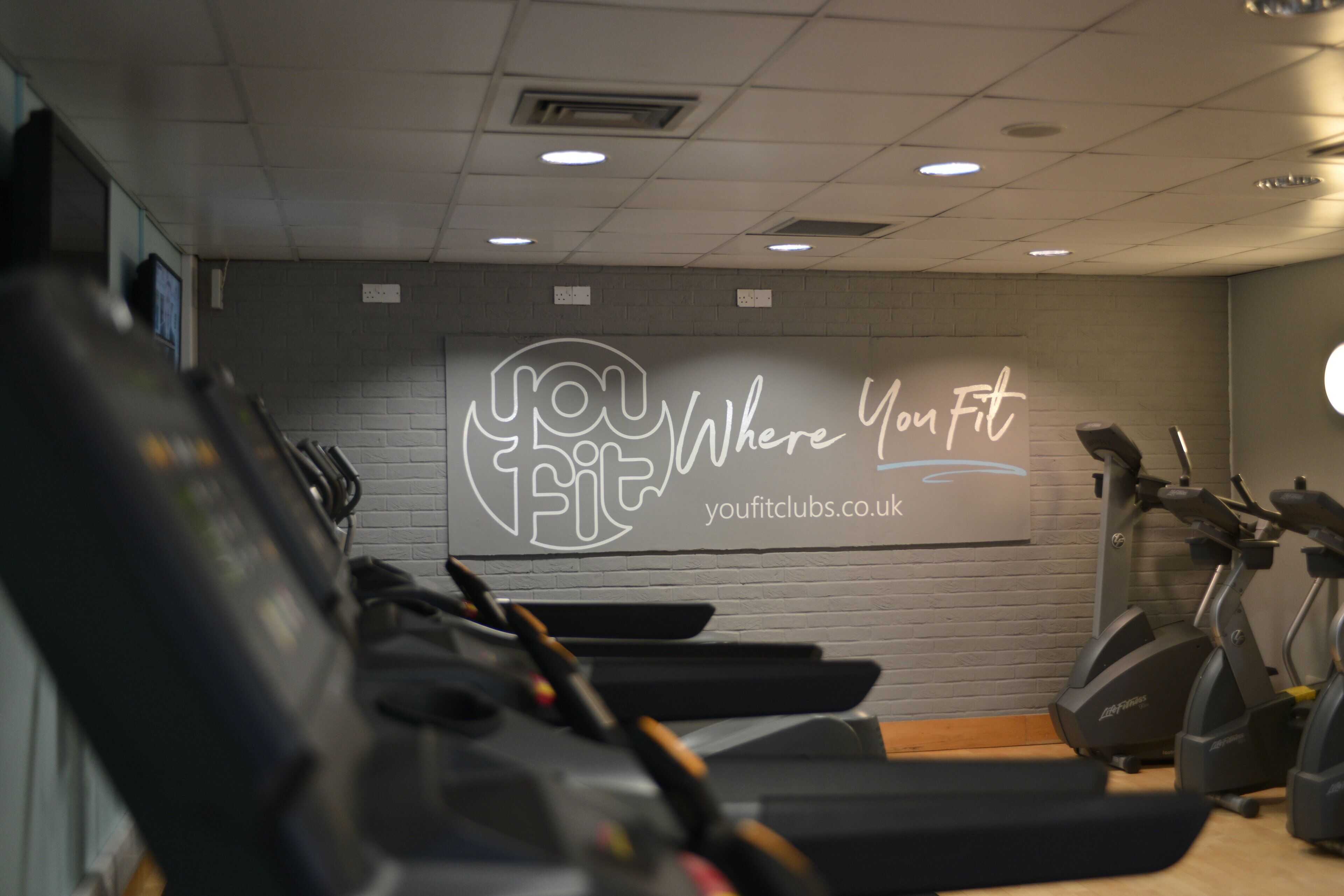 A gym with treadmills and stationary bikes, a motivational wall with the text 'Where You Fit' and a website address.