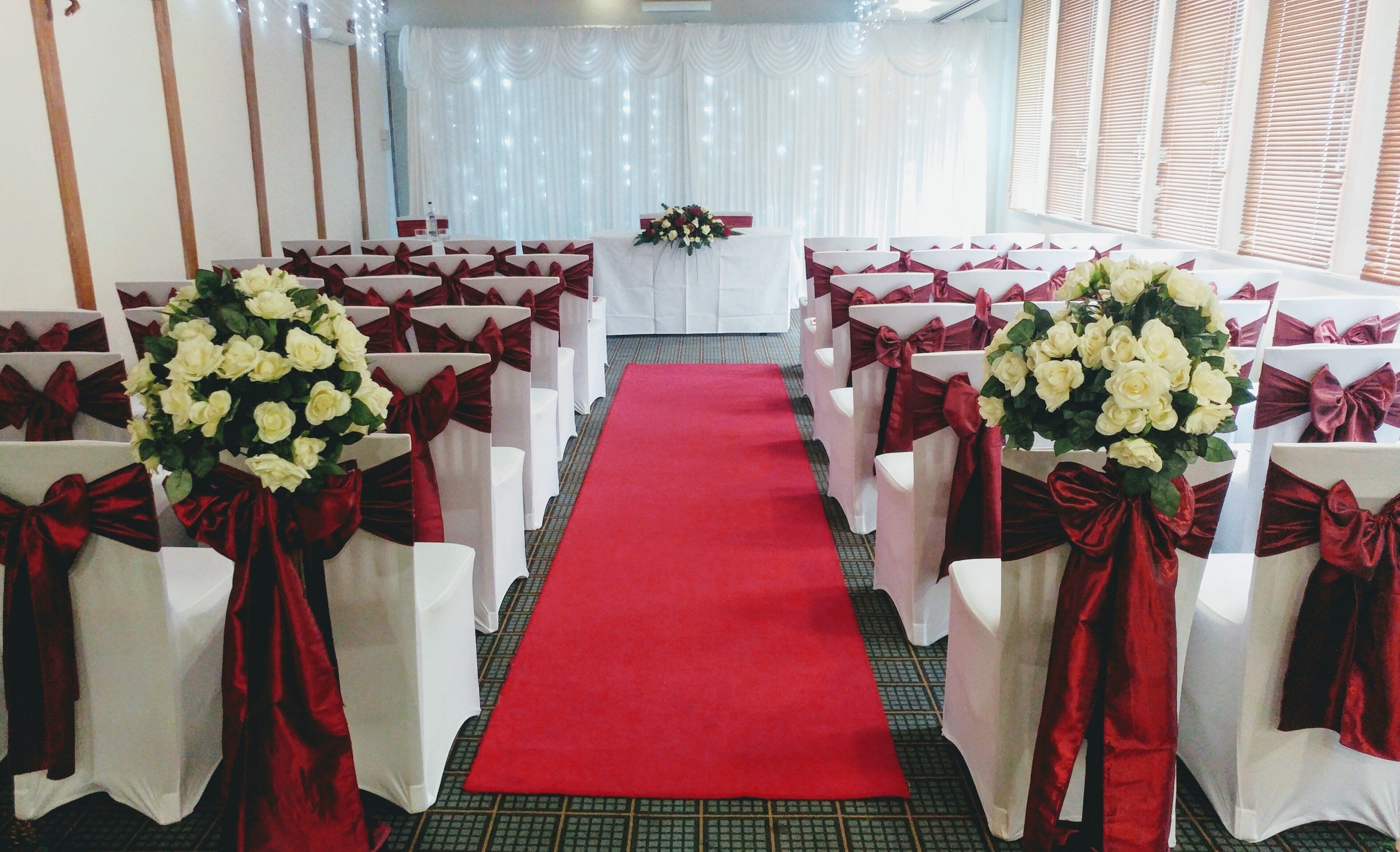 A red carpet aisle leading to a white table with floral arrangements, flanked by rows of white chairs with red sashes.