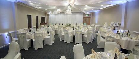 Un salón de banquetes con mesas redondas dispuestas para comer, cortinas blancas y una araña de cristal.