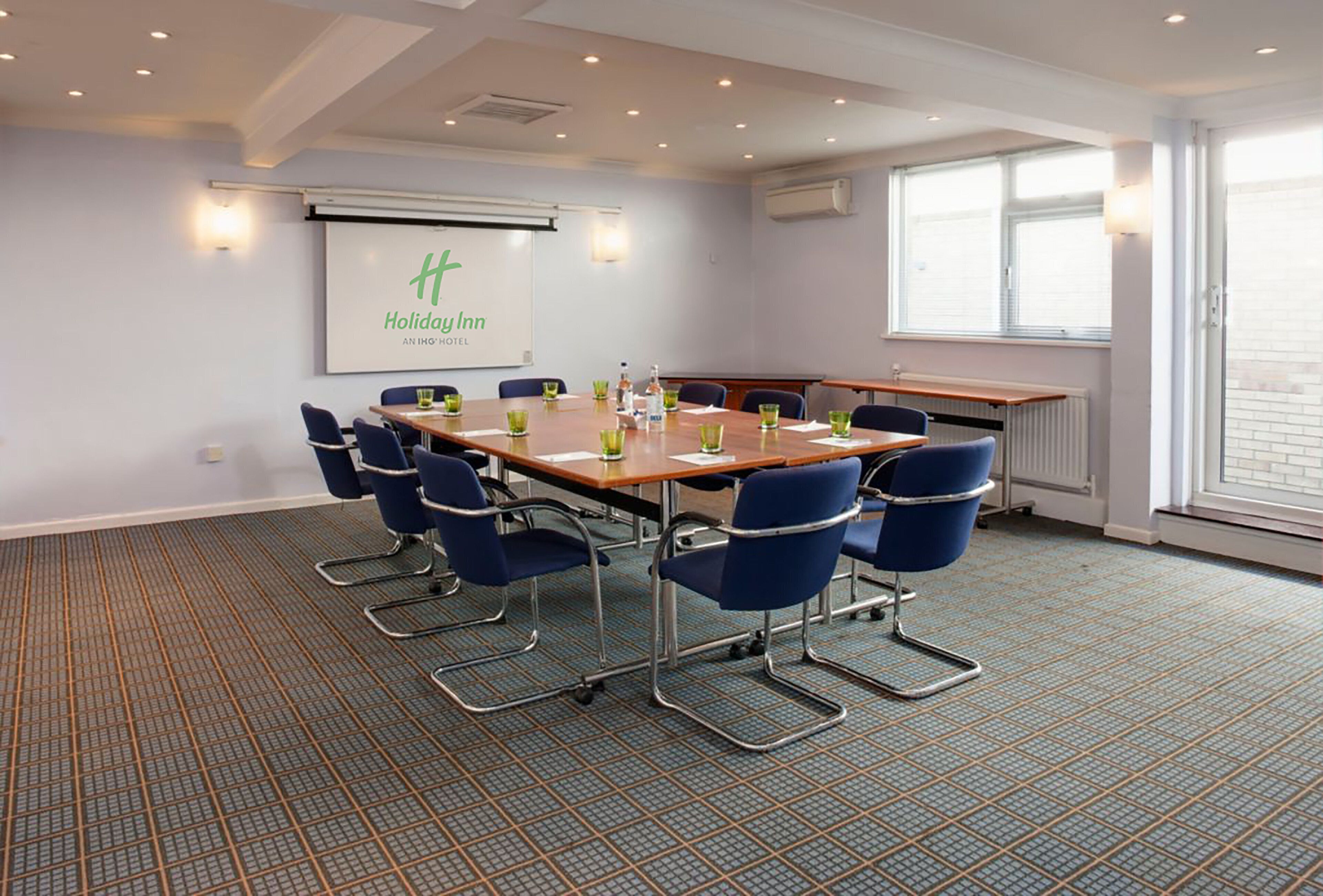 A conference room with a large table, blue chairs, a projector screen, and a window with blinds.