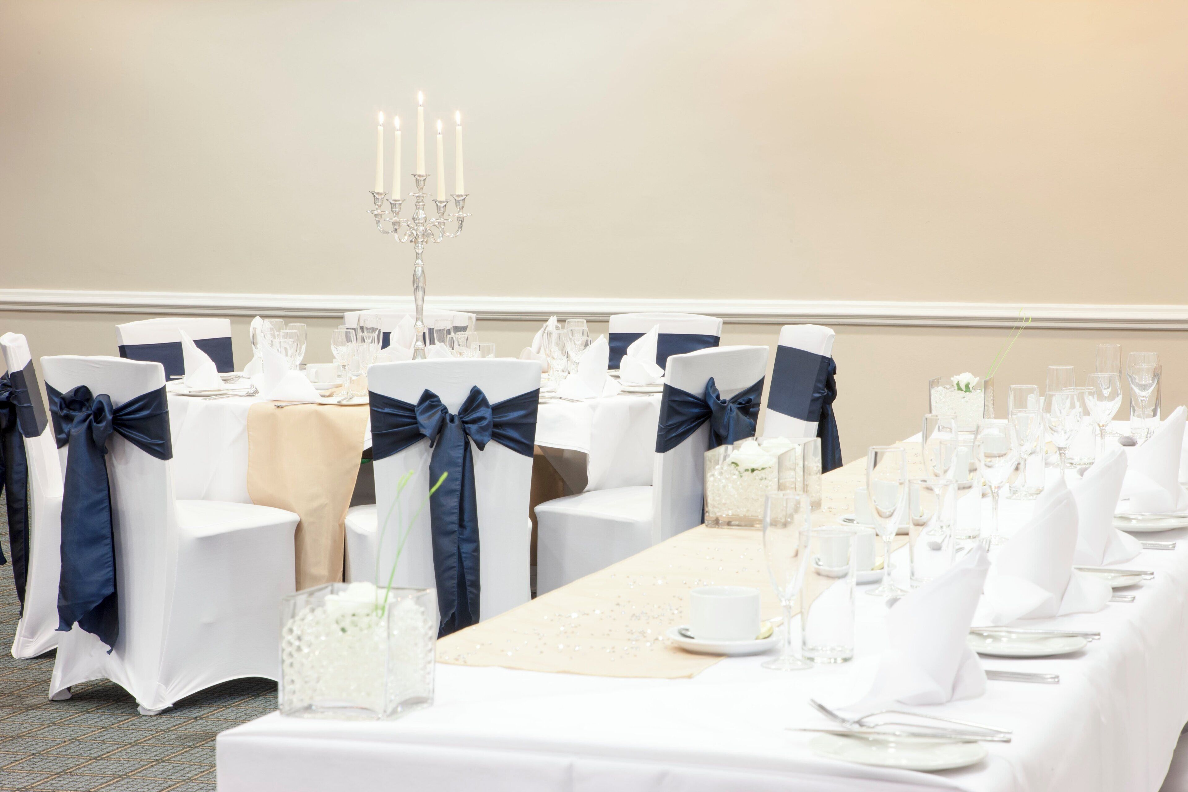 A banquet hall with round tables set for a formal event, featuring white tablecloths, neatly arranged glassware, and chairs with navy blue sashes.