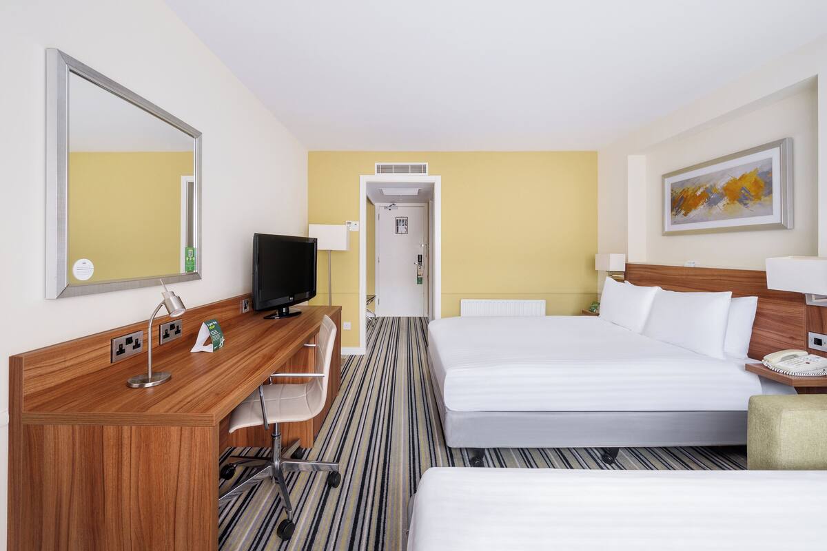 A hotel room with a bed, a desk with a TV, a chair, a bedside table with a lamp, and a painting on the wall.