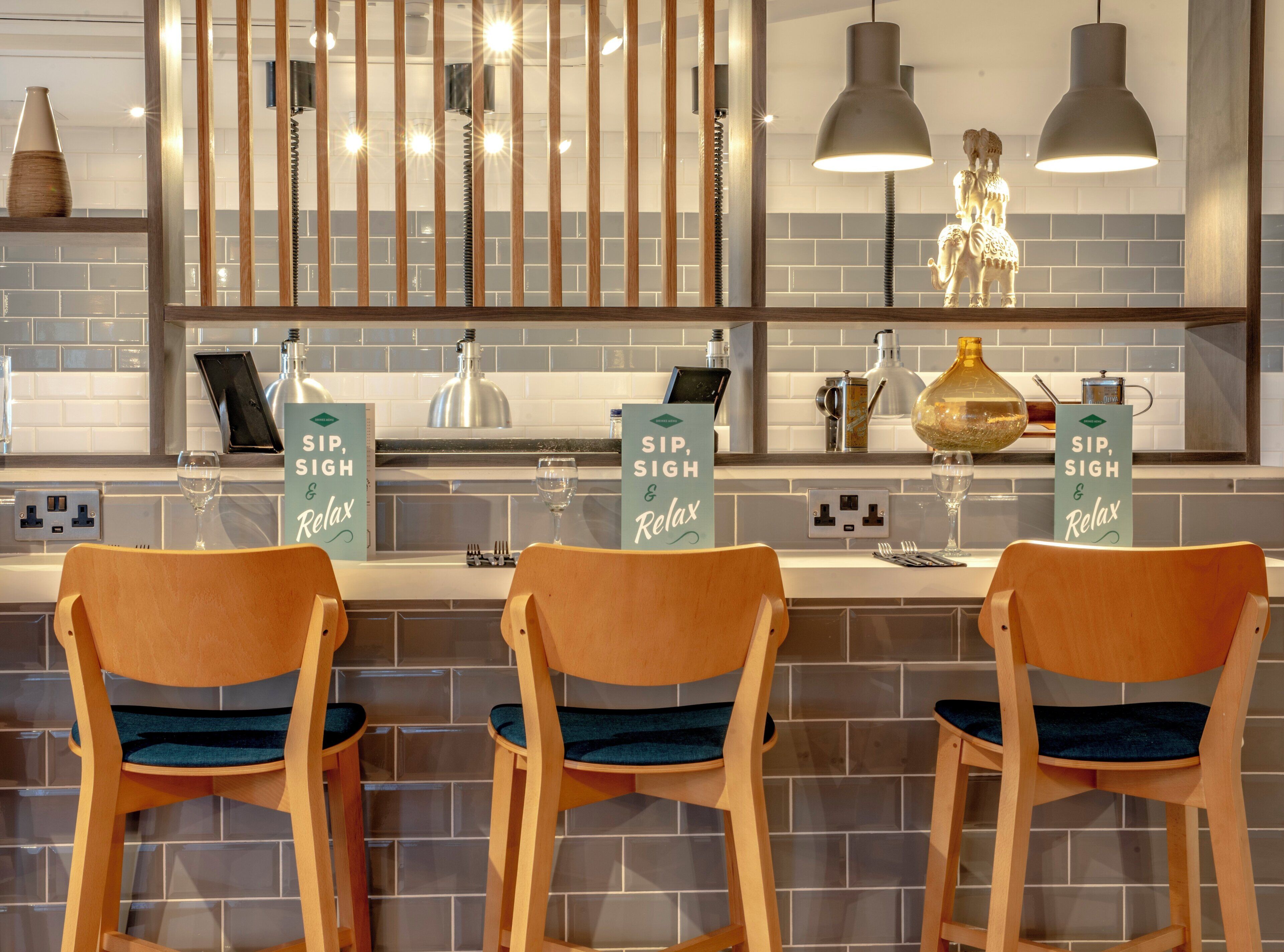 A bar area with three wooden chairs, a countertop with bar tools, and two signs that read 'SIP, SIGH & Relax'.