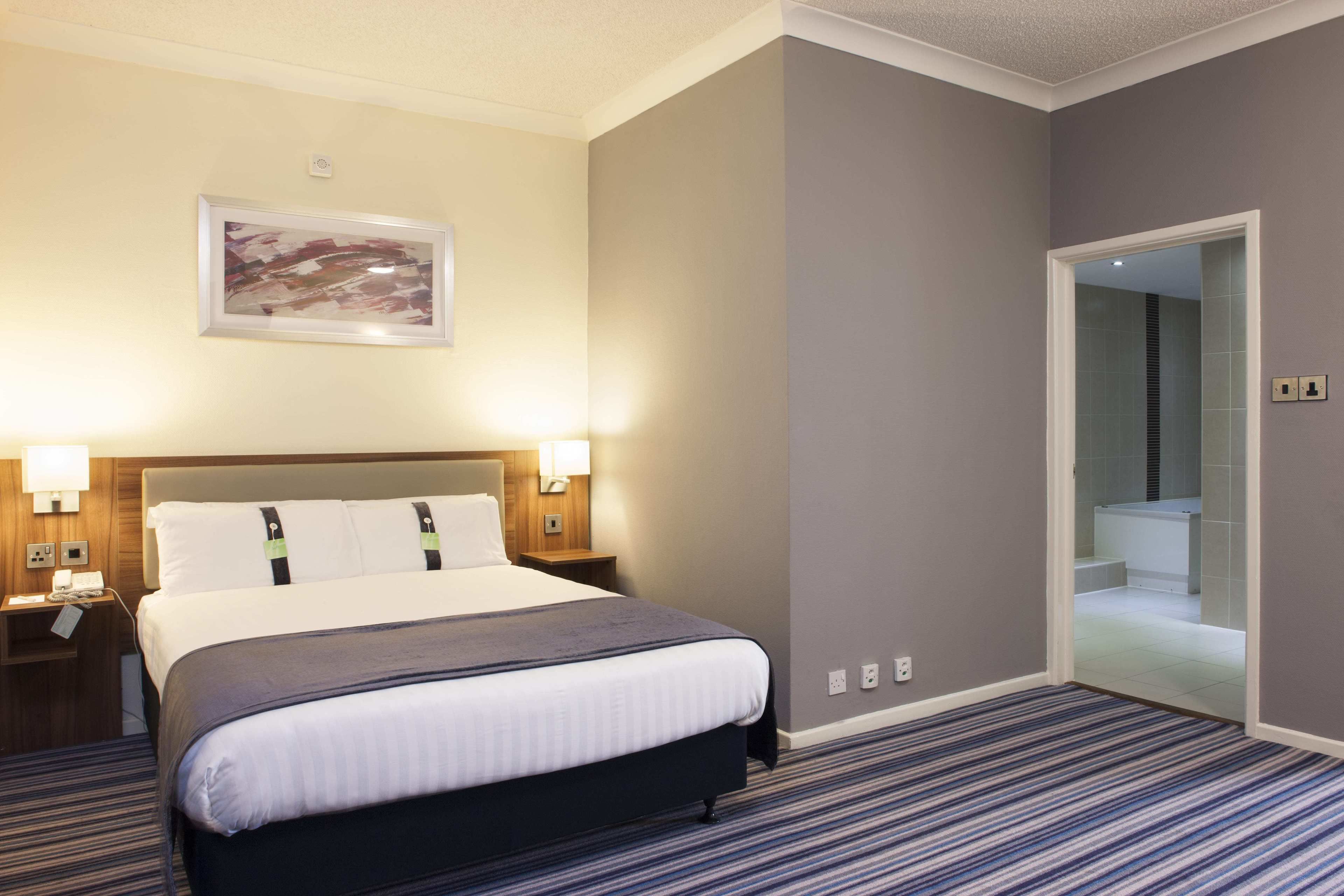 A hotel room with a large bed, bedside lamps, a framed artwork on the wall, and a bathroom visible through an open door.