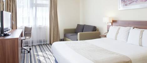 Quarto de hotel com cama, escrivaninha com computador, cadeira, sofá e um quadro na parede.