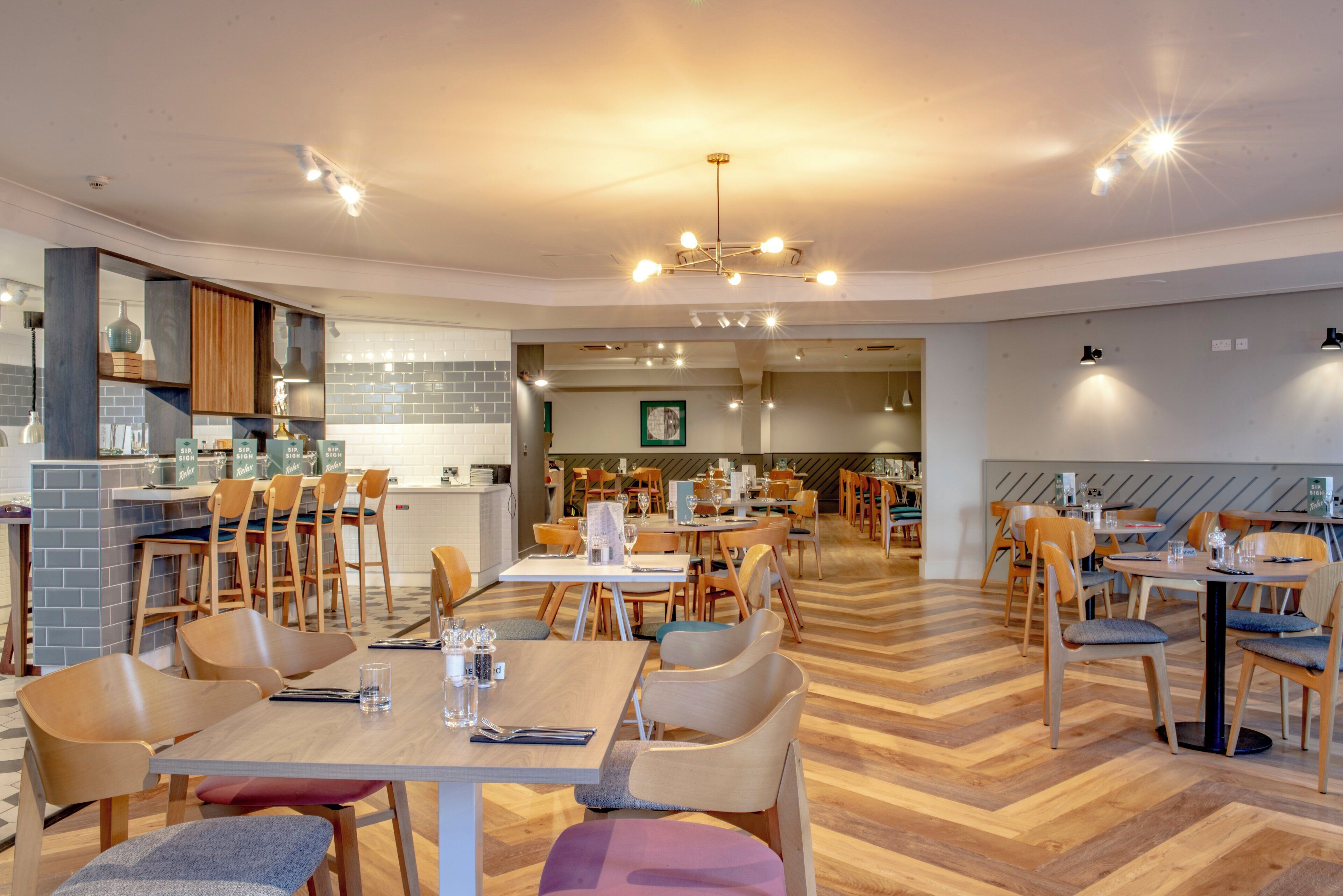 A modern dining area with wooden tables and chairs, a kitchen area with a brick backsplash, and a visible ceiling light fixture.