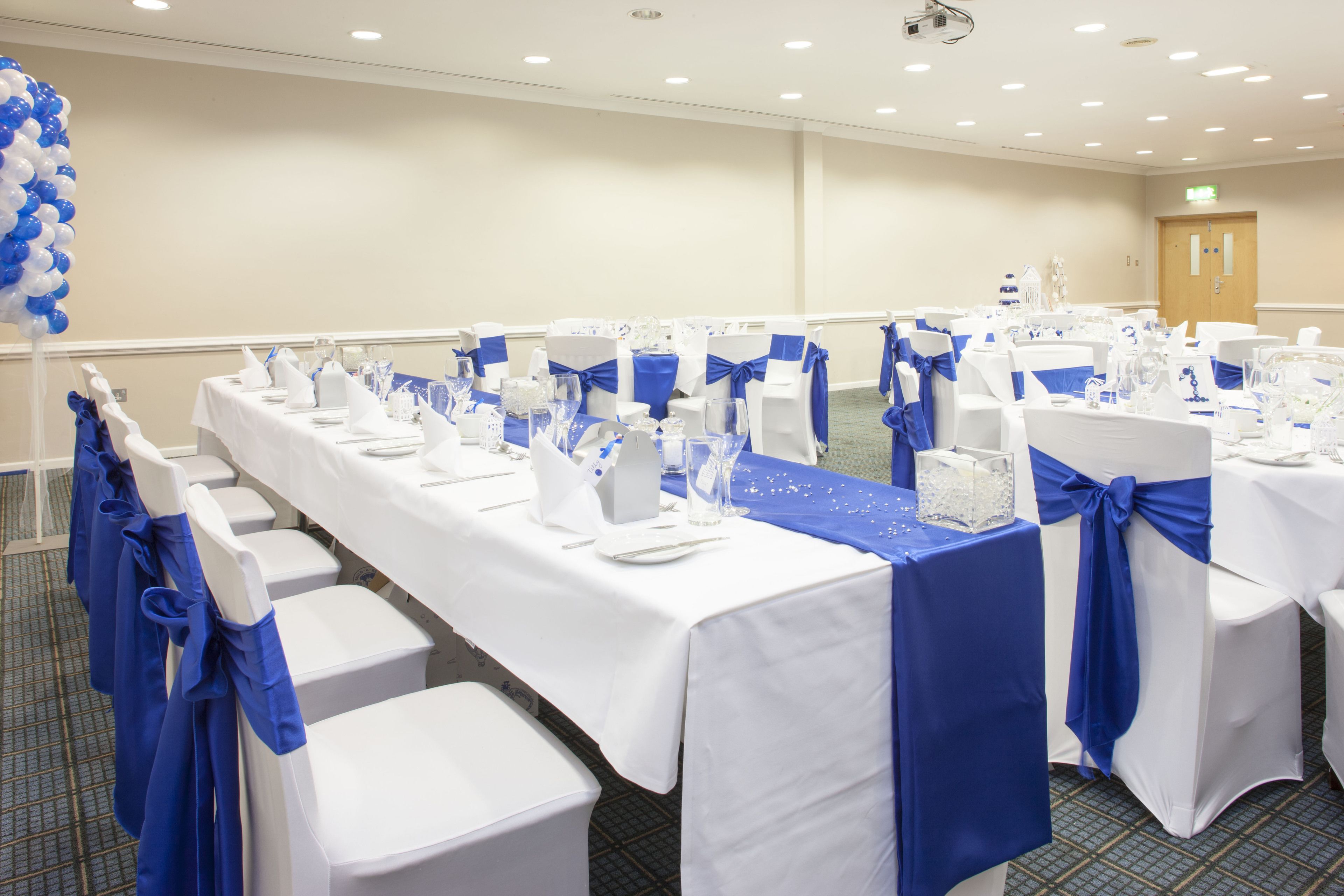 A banquet hall with tables set for a formal event, featuring white tablecloths and blue sashes.