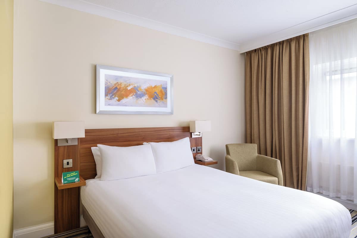 A hotel room with a large bed, a wooden headboard, two bedside lamps, a chair, a painting on the wall, and a window with curtains.