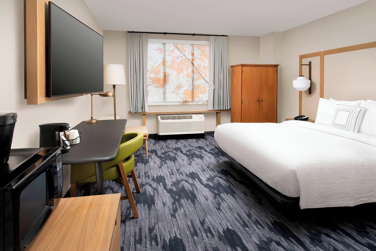 A hotel room with a bed, a desk with a computer, a chair, a TV, a wardrobe, and a window with curtains.