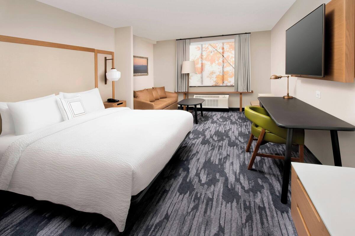 Ein Hotelzimmer mit einem großen Bett, einem Schreibtisch, einem Sessel, einem Fernseher und einem Fenster mit Vorhängen.