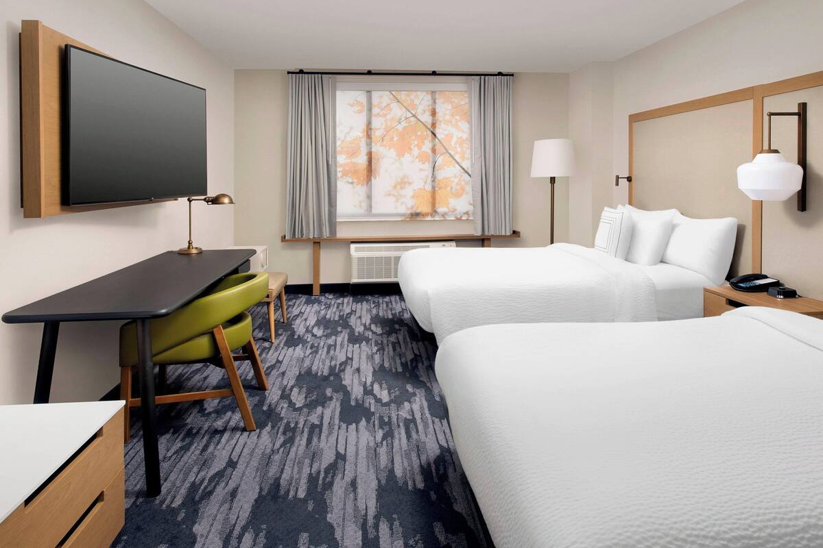 Ein Hotelzimmer mit zwei Betten, einem Schreibtisch mit Stuhl, einem Fernseher, einem Fenster mit Vorhängen und einer Lampe.