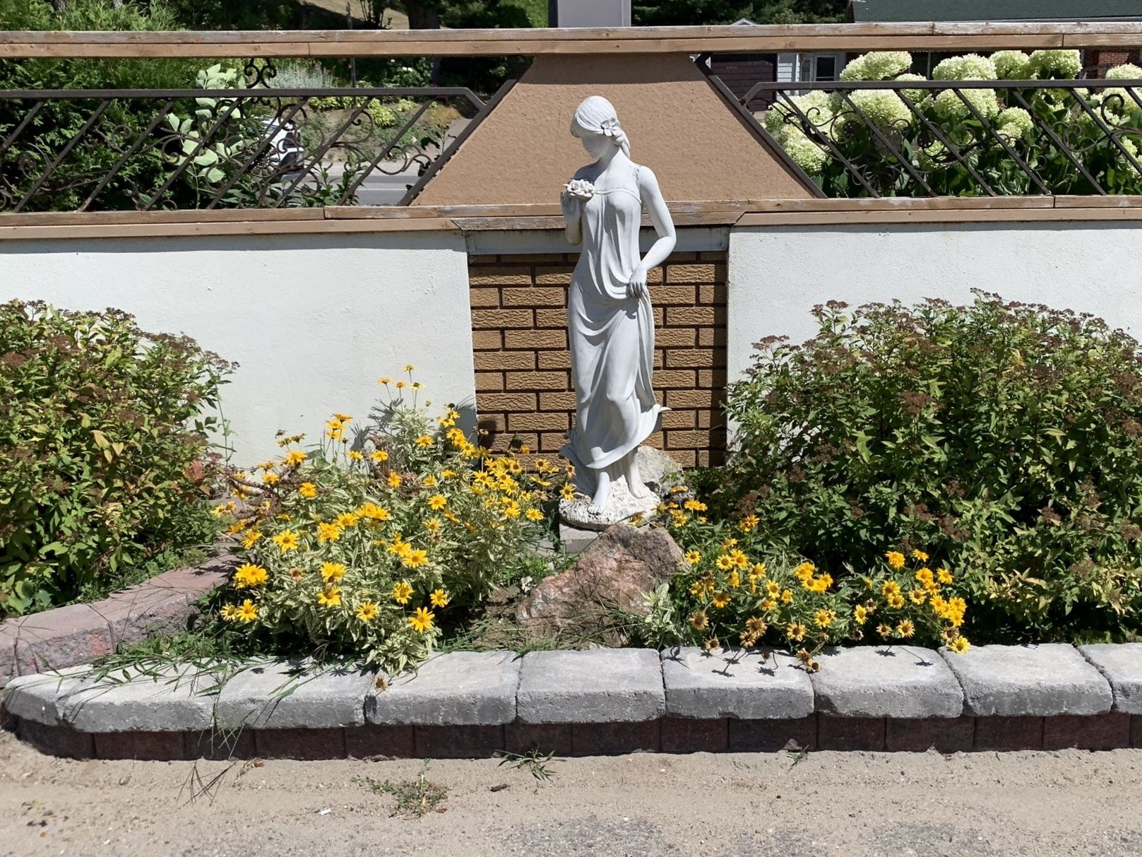 A statue of a woman holding a flower, surrounded by yellow flowers and greenery.