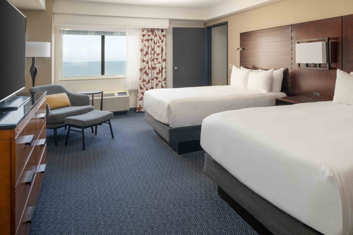A hotel room with two beds, a desk, a chair, a TV, and a view of the ocean.