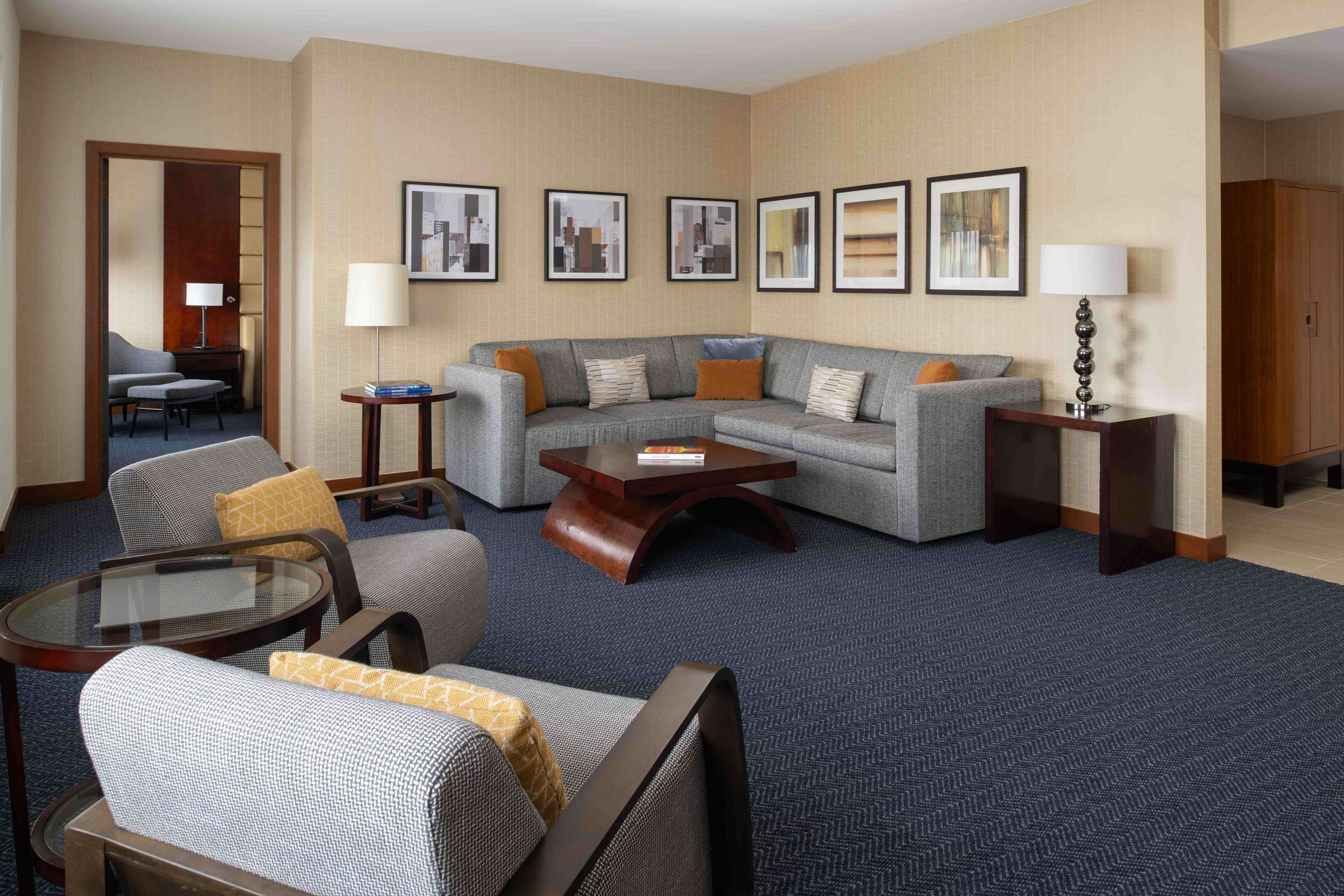 A hotel room with a sectional sofa, a glass coffee table, a small round table, and a chair. There are framed pictures on the wall and a lamp on a side table.