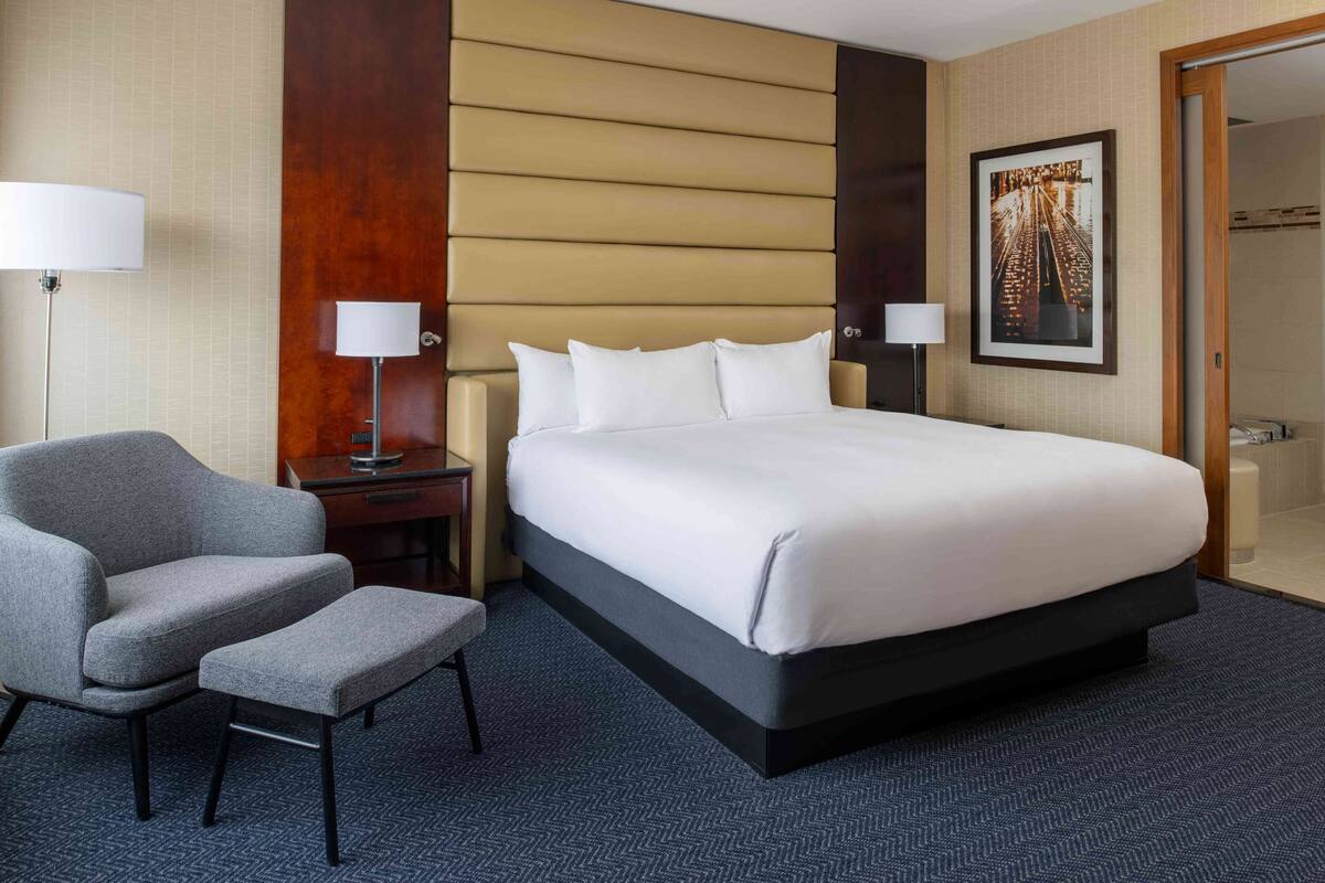 A hotel room with a large bed, a grey armchair, a small table, and two lamps.