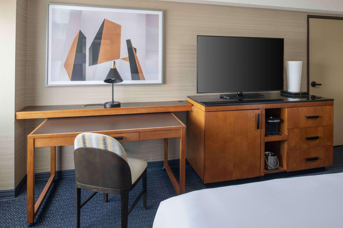 A hotel room with a wooden desk, a chair, a flat-screen TV, a cabinet with drawers, and a bed.