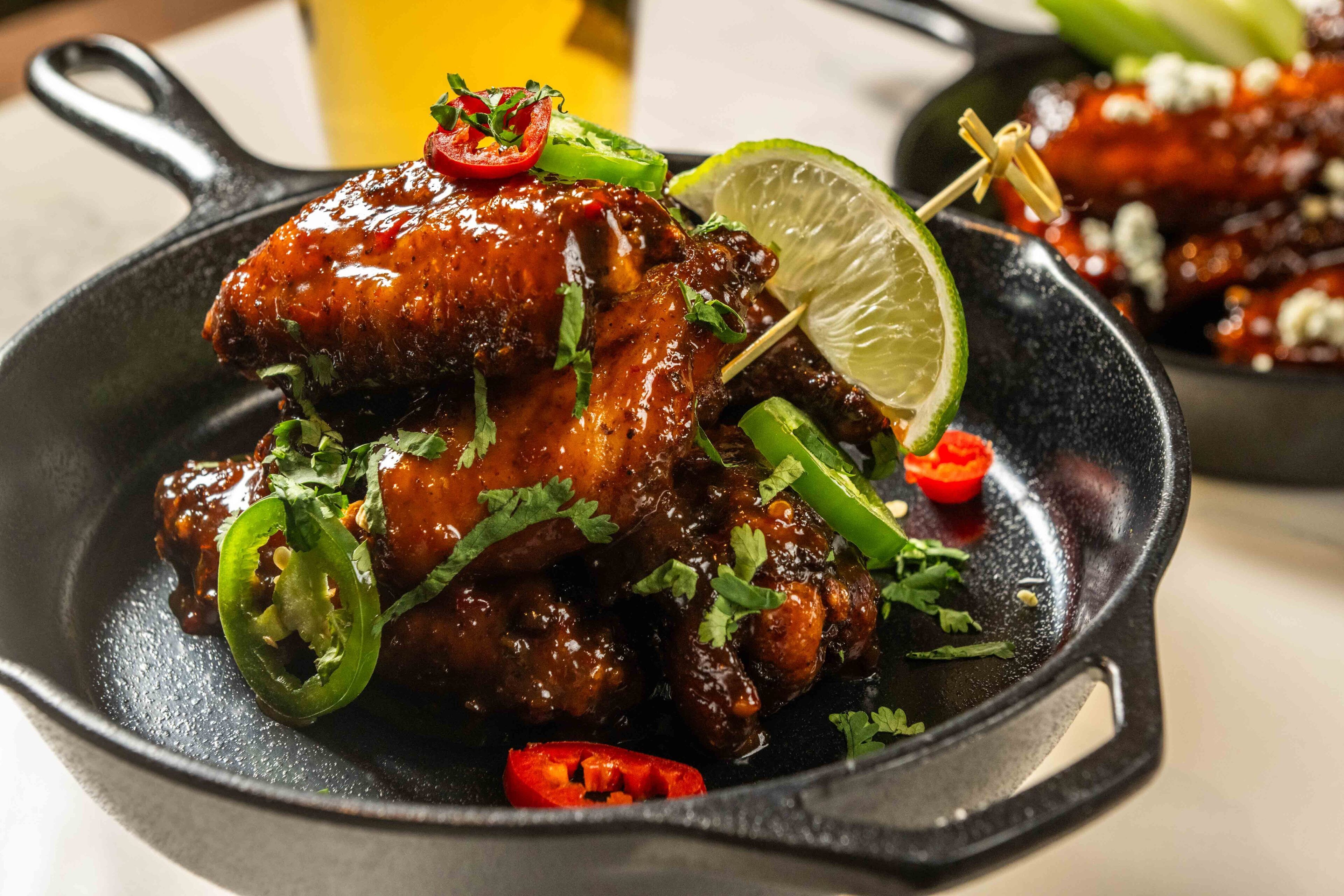 A skillet with grilled chicken wings garnished with lime, cilantro, and sliced jalapeños.