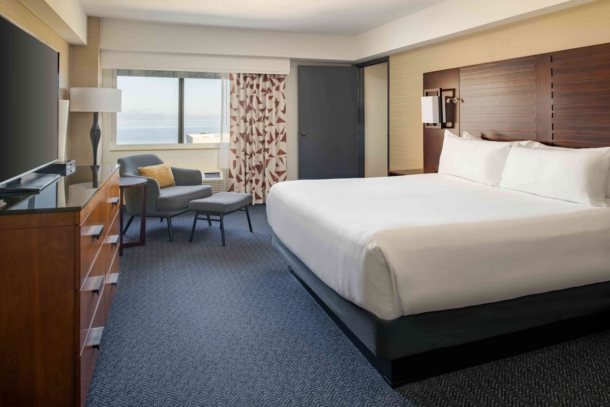 A hotel room with a large bed, a desk with a lamp, a chair, and a view of the sea through the window.