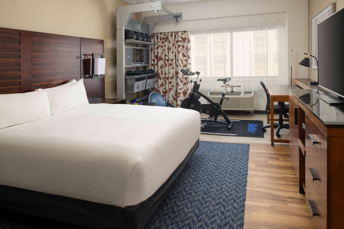A hotel room with a large bed, a desk, a chair, a TV, a treadmill, and a small kitchenette.