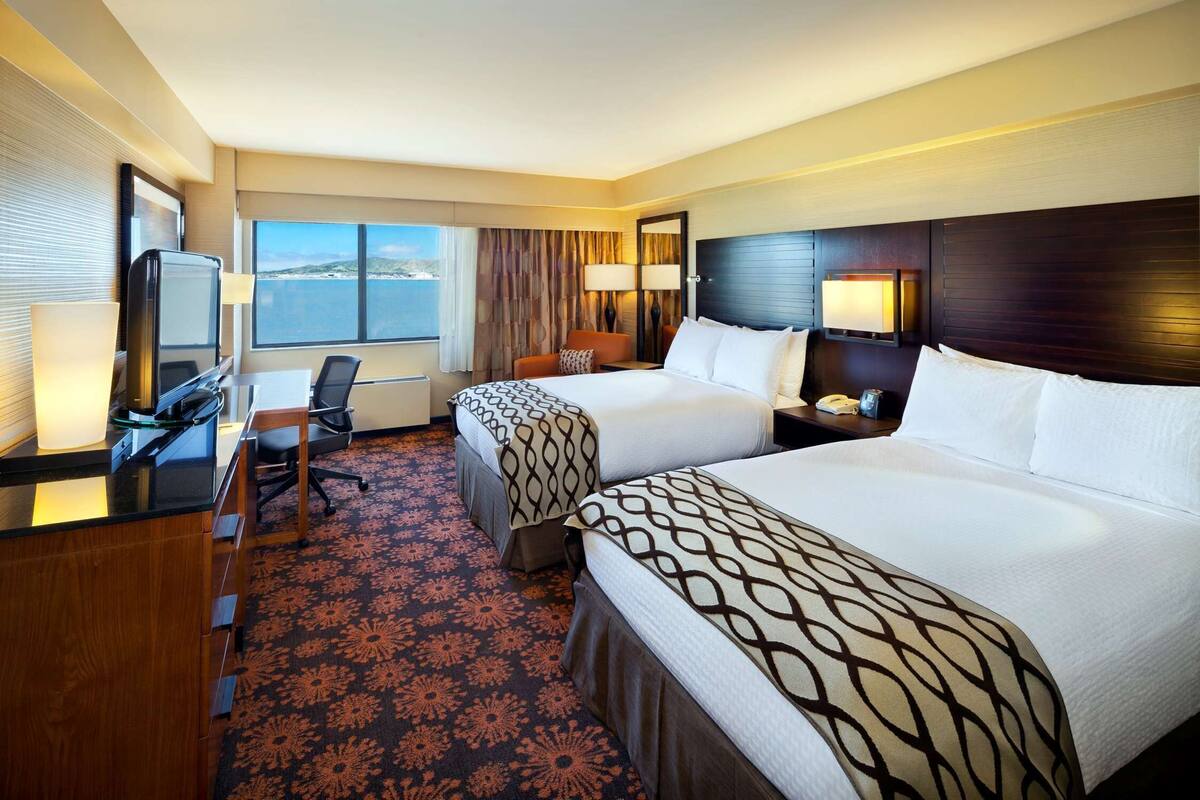 A hotel room with two beds, a desk, a chair, a TV, and a large window with a view.