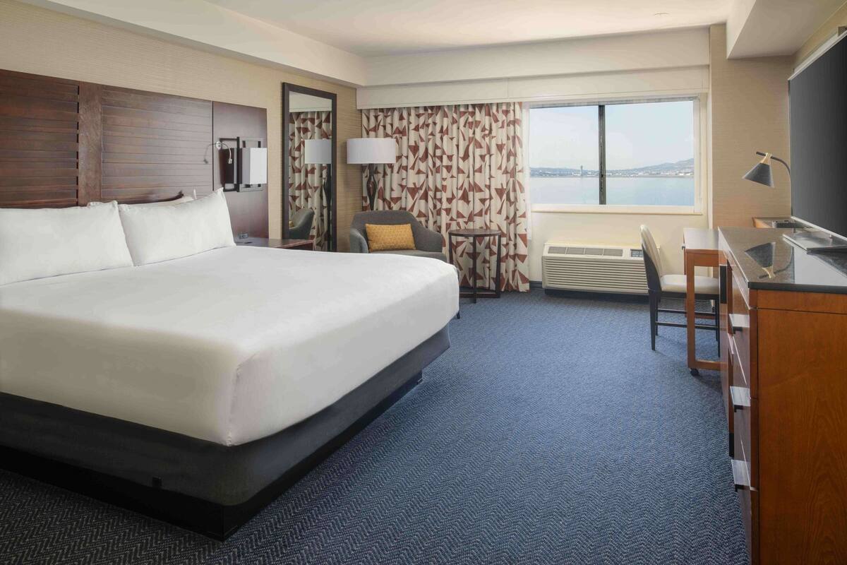 A hotel room with a large bed, a desk, a chair, a lamp, and a view of the city through the window.