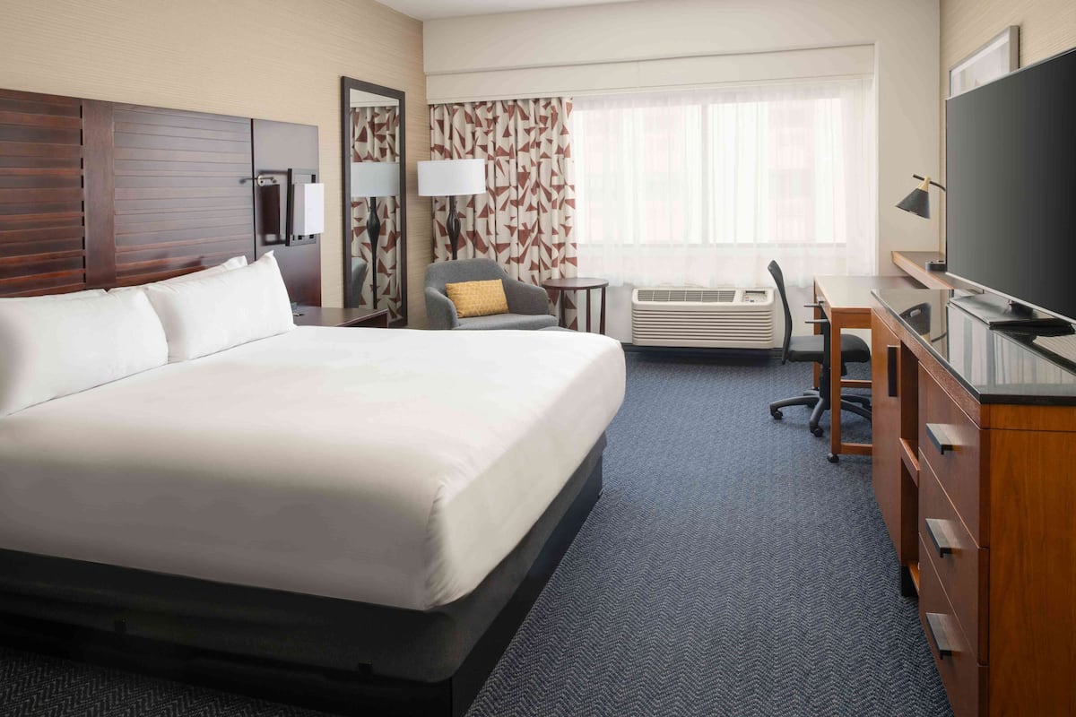 A hotel room with a large bed, a desk with a computer, a chair, an air conditioning unit, and a television.