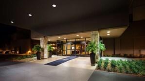 A hotel entrance with a dark facade, glass doors, and a welcoming outdoor area with potted plants and a carpeted path.