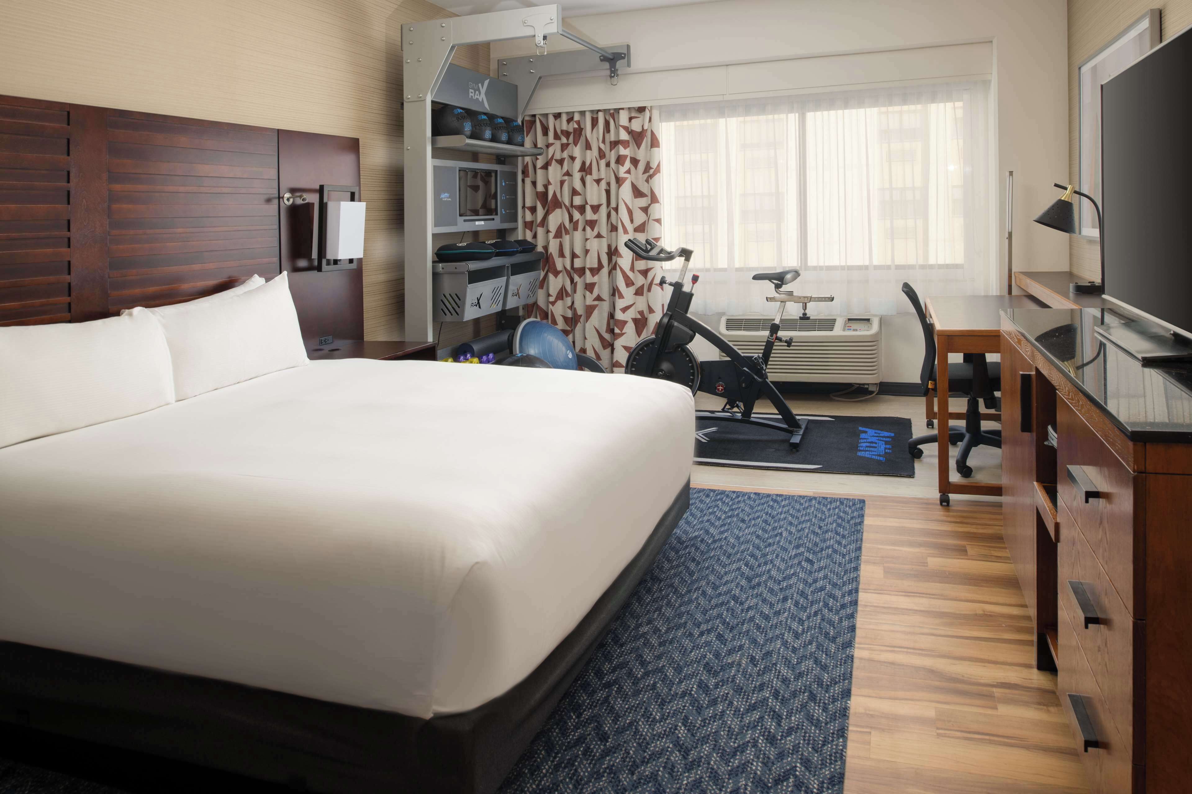 Una habitación de hotel con una cama grande, un escritorio con una computadora, un televisor y una bicicleta estática.