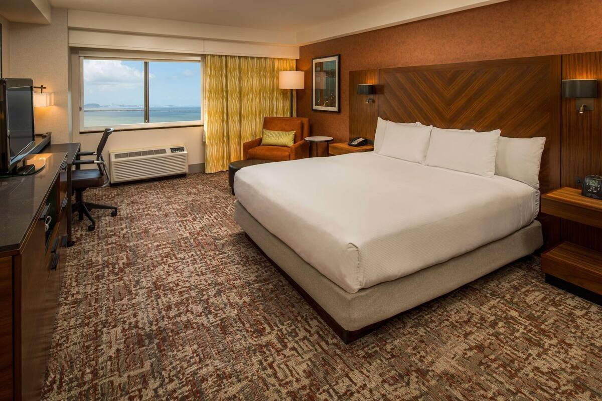 A hotel room with a large bed, a desk with a computer, a chair, a small table, and a view of the sea.