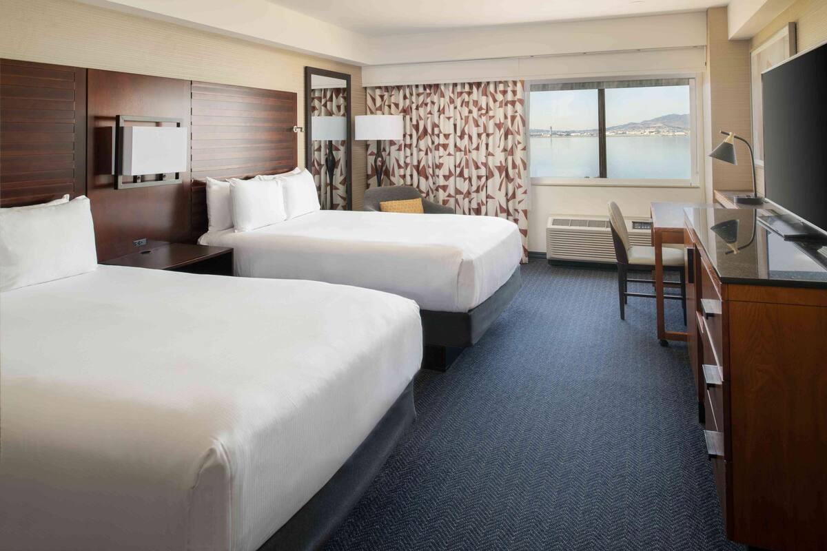 A hotel room with two beds, a desk, a chair, a TV, and a large window with a view of the sea.