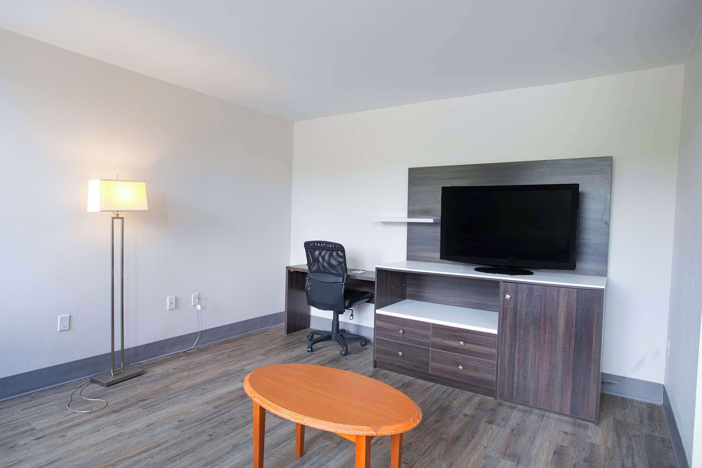A modern hotel room with a wooden floor, a desk with a chair, a flat-screen TV mounted on a wooden cabinet, and a floor lamp.