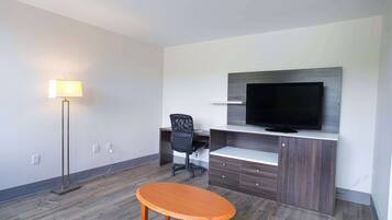 A modern hotel room with a wooden floor, a desk with a chair, a flat-screen TV mounted on a wooden cabinet, and a floor lamp.
