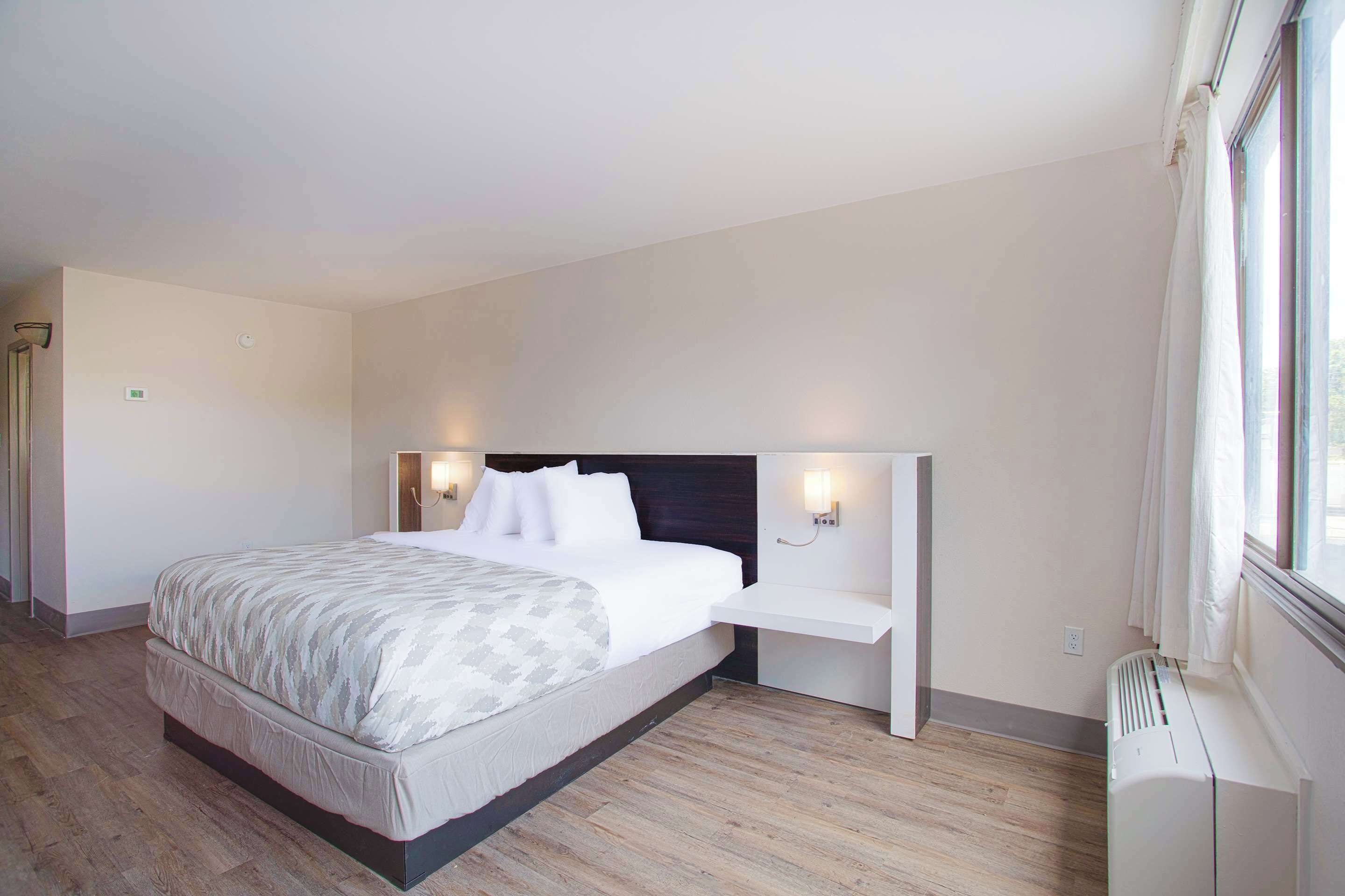 A modern hotel room with a large bed, bedside tables with lamps, a built-in heater, and a window with curtains.