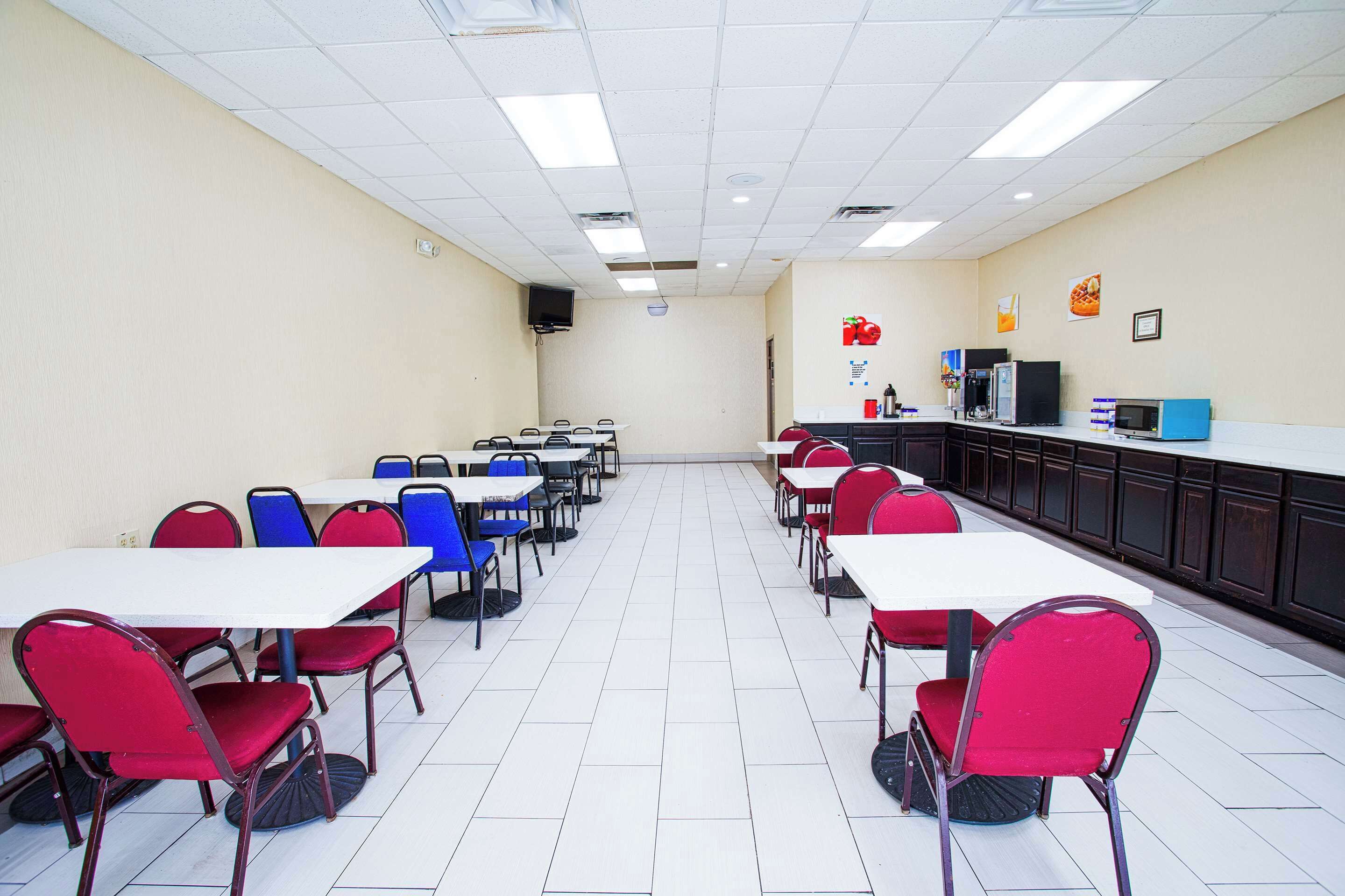 A cafeteria with white tables and chairs, a kitchen area, and a tiled floor.