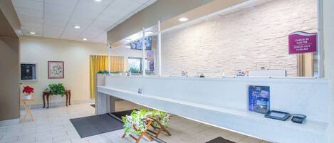 A reception area with a white reception desk, a check-in/out sign, a digital kiosk, and a small plant arrangement.