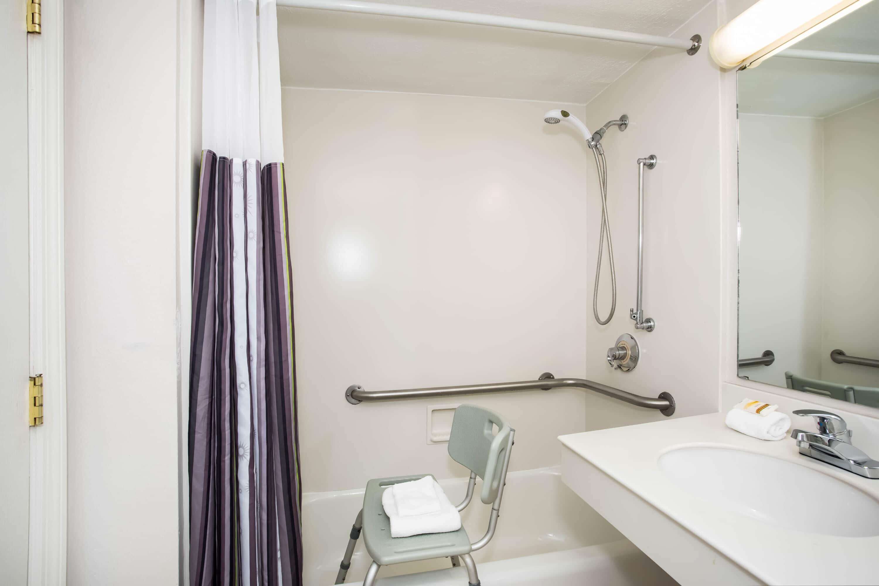 A bathroom with a shower, a sink, a chair, and a handrail.